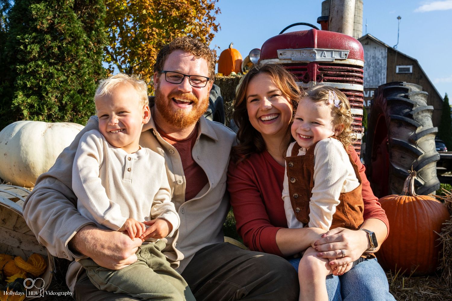 Family sitting together by a red tractor surrounded by pumpkins during fall harvest.
