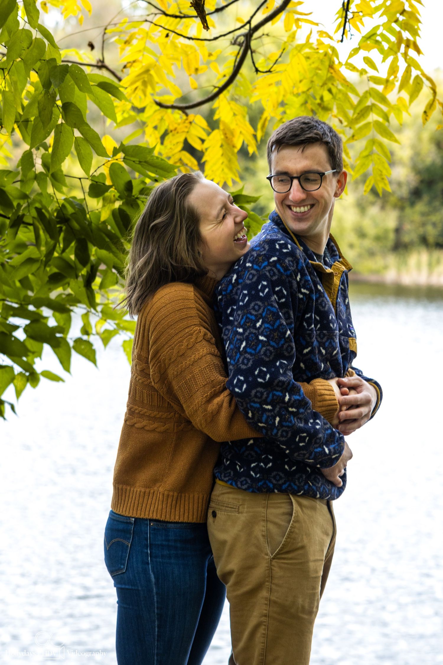 Couple embracing under yellow fall foliage near a lake.