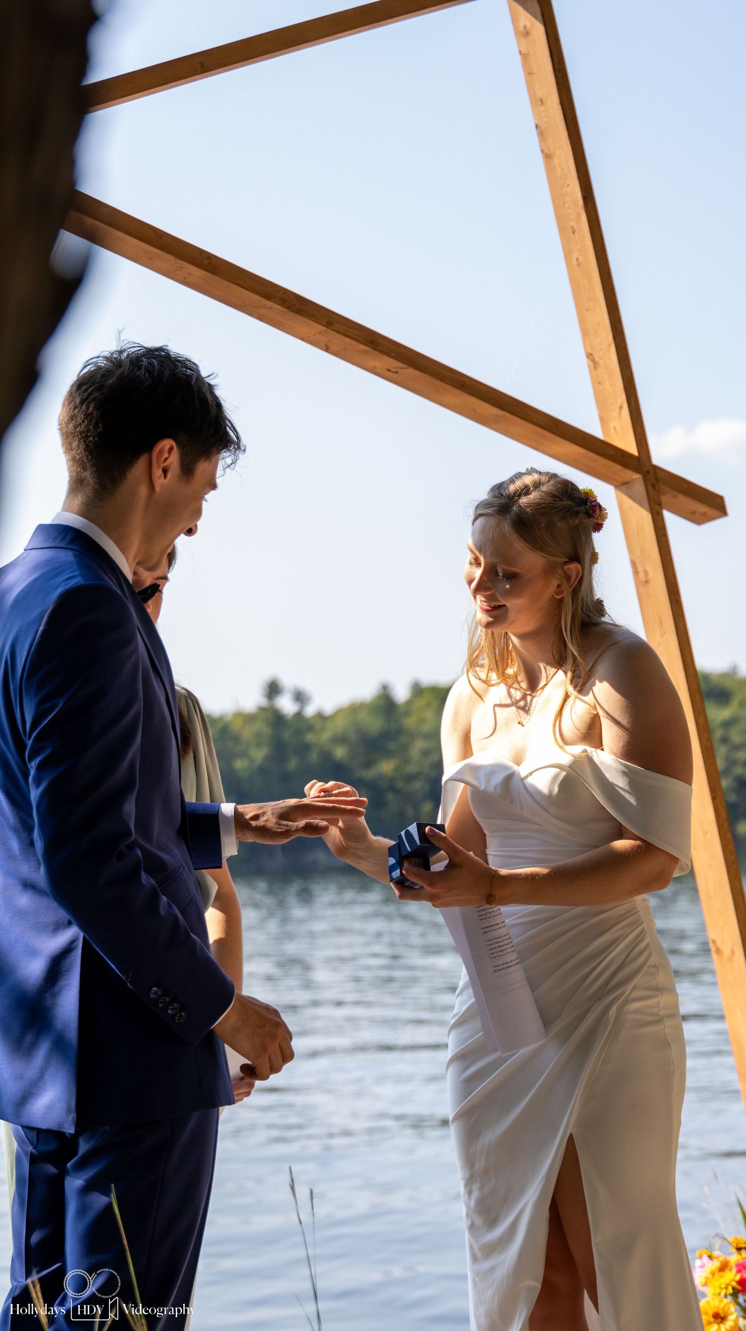 A couple exchanges rings during a lakeside wedding ceremony under a wooden arch.