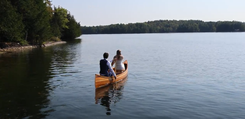 Two people paddle in wooden canoe across serene lake surrounded by trees.