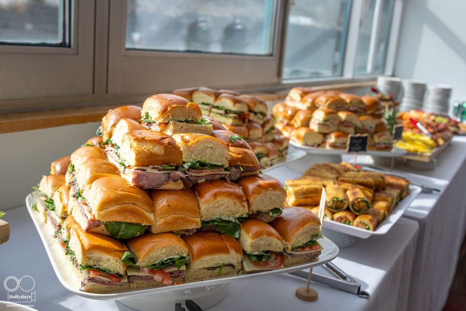 Rows of mini sandwiches and appetizers arranged on white plates at a catering event by a window.