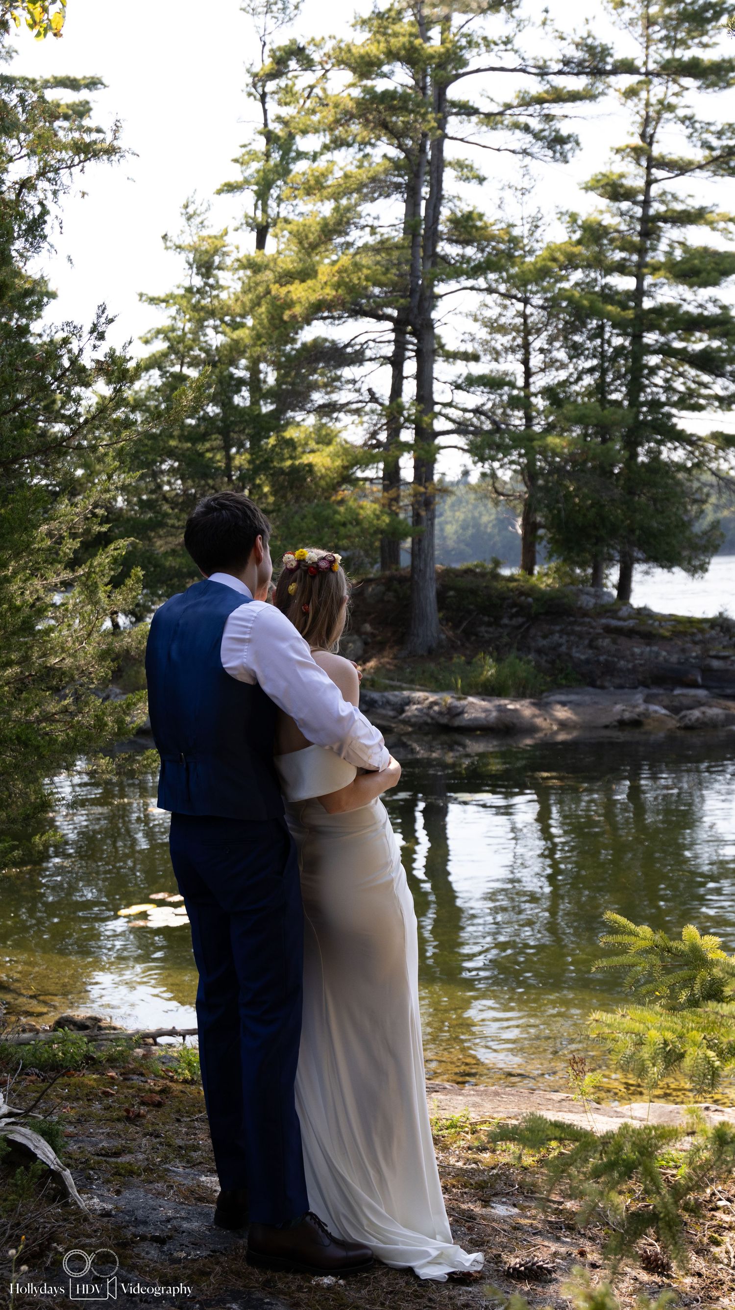 A couple shares an intimate moment overlooking a serene lake surrounded by tall pine trees at sunset.
