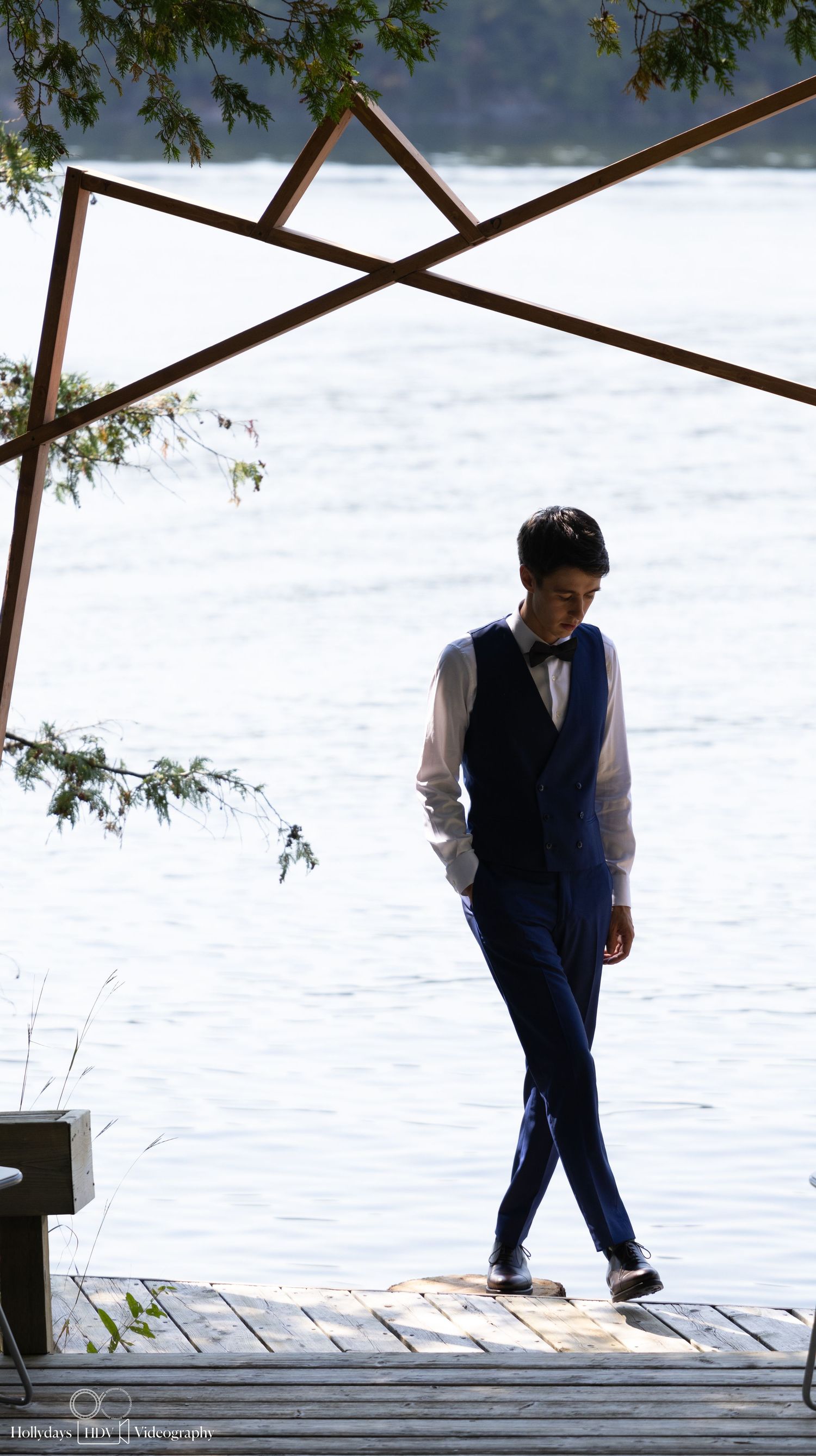 Figure in formal attire stands contemplatively on wooden dock under geometric arch structure.