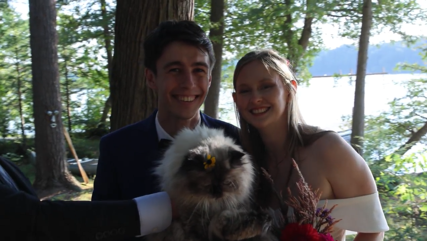 Wedding couple poses with fluffy dog outdoors by lakeside trees.