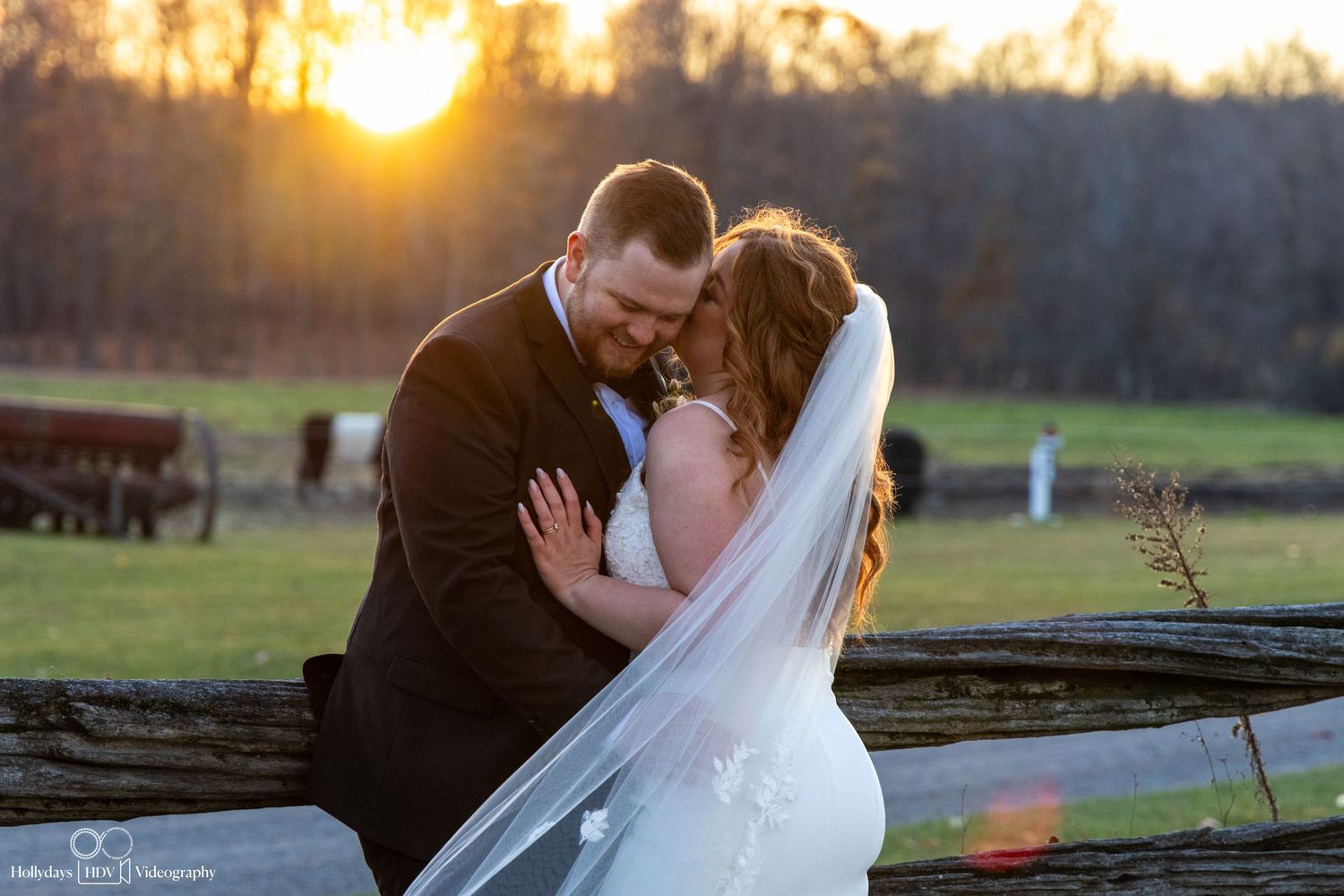 A couple in wedding attire embraces at sunset on a farm with a flowing veil and golden light in the background.