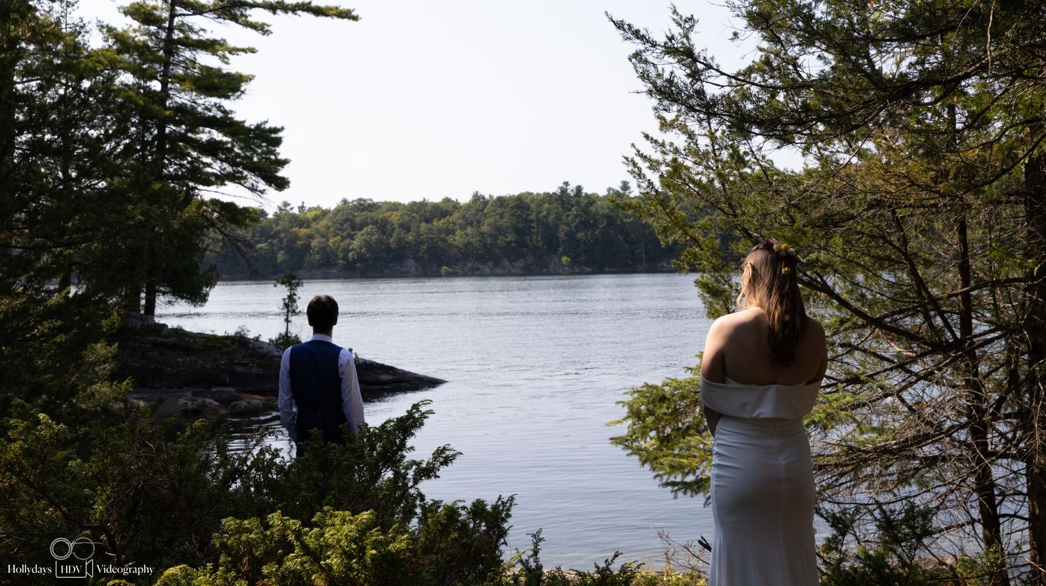 Two people stand at lakeside viewpoint surrounded by pine trees.