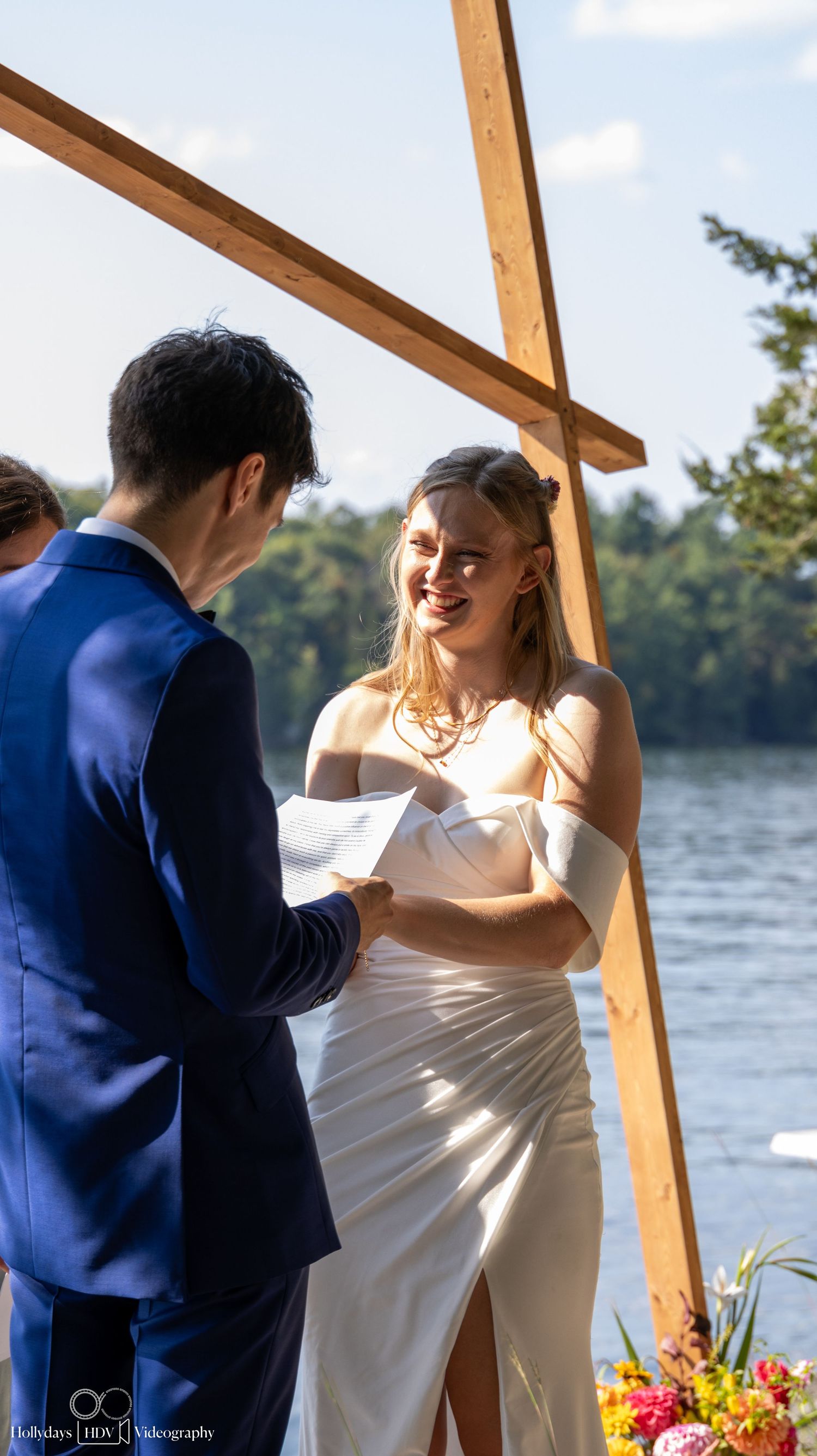 A couple reads handwritten vows during their lakeside wedding ceremony under wooden beams.