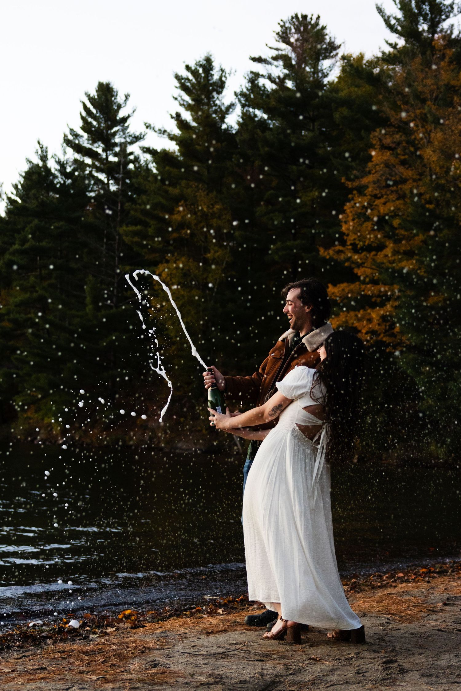 Person in a white dress creates magical water splashes during sunset by a lake with pine trees.