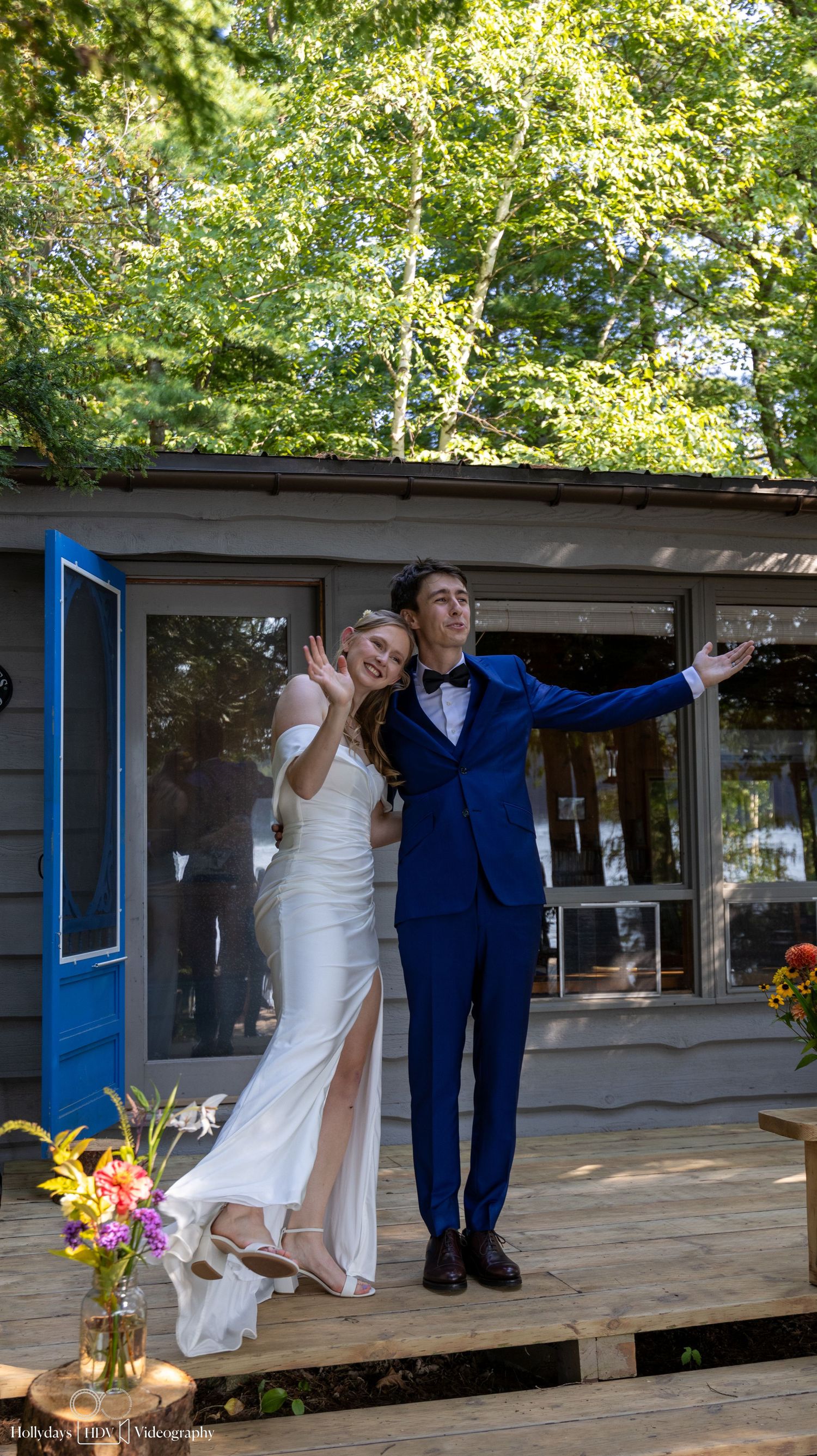 A couple in wedding attire poses playfully on a rustic porch with string lights and flowers.