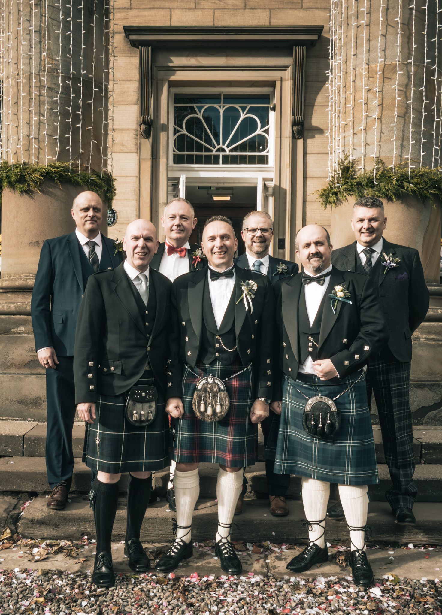 Seven men in Scottish kilts and formal attire pose on stone steps at a traditional wedding ceremony.