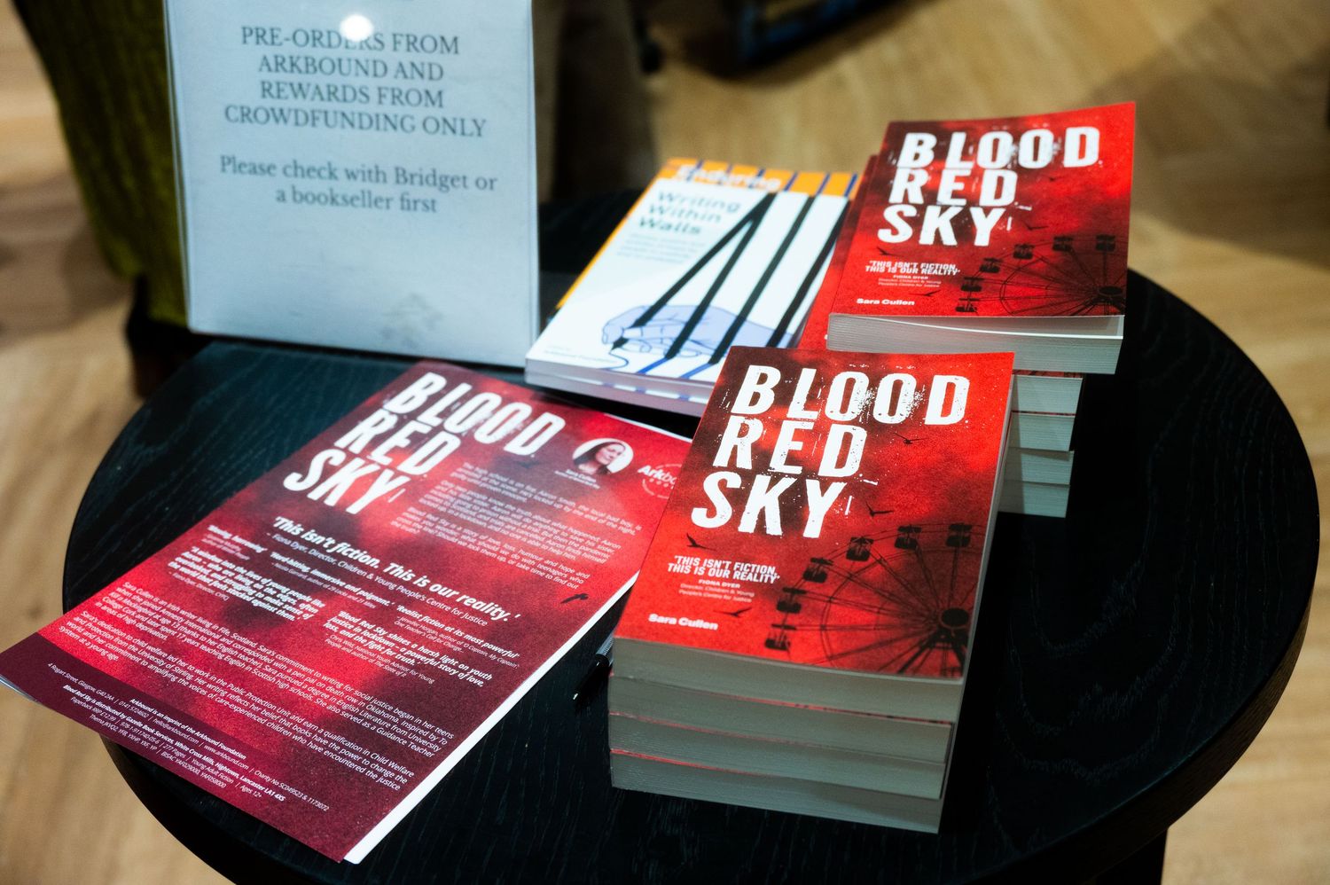 Stack of Blood Red Sky books displayed on a table at a book signing event with promotional flyers.