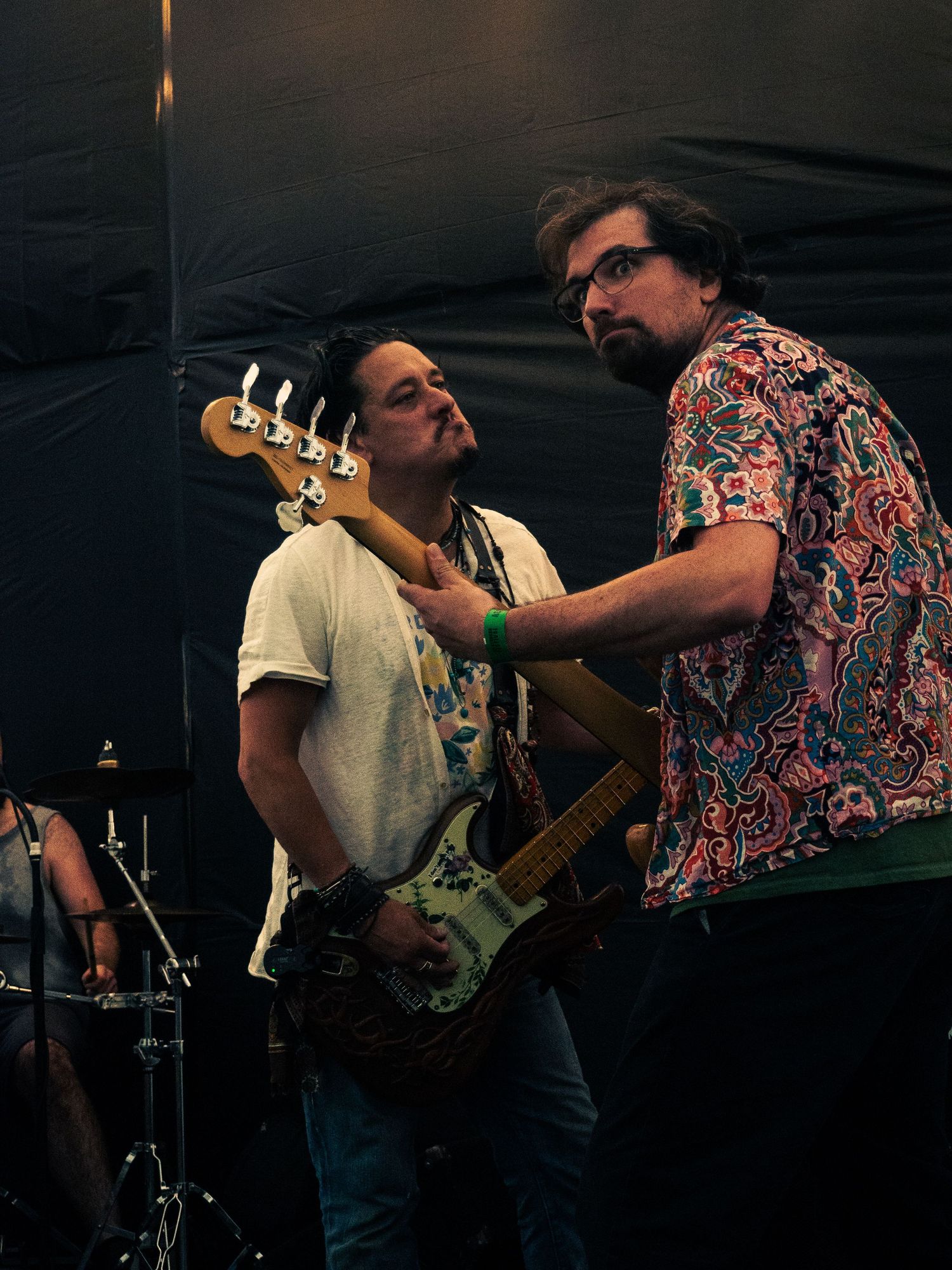 Two musicians interact on stage, one holding a bass guitar while wearing a colorful patterned shirt.
