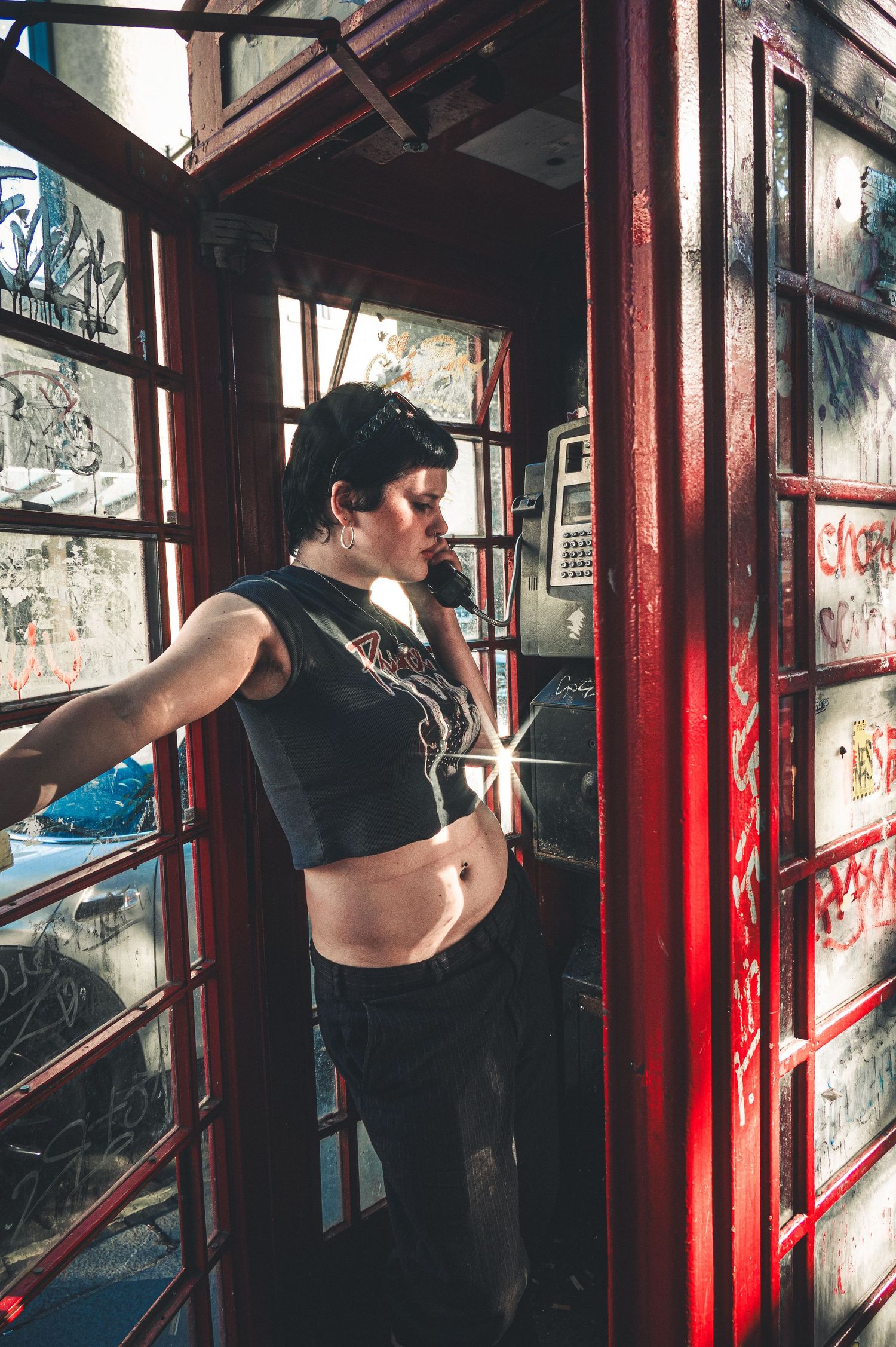 A young woman in a crop top poses in a graffiti-covered red phone booth while talking on a vintage payphone.