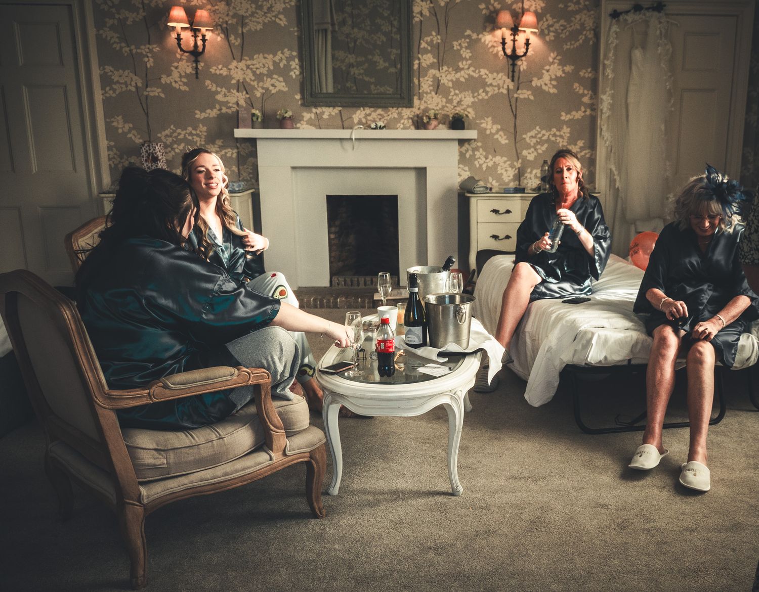 Three women relaxing in an elegant room with champagne, seated on vintage chairs and a sofa near a fireplace.