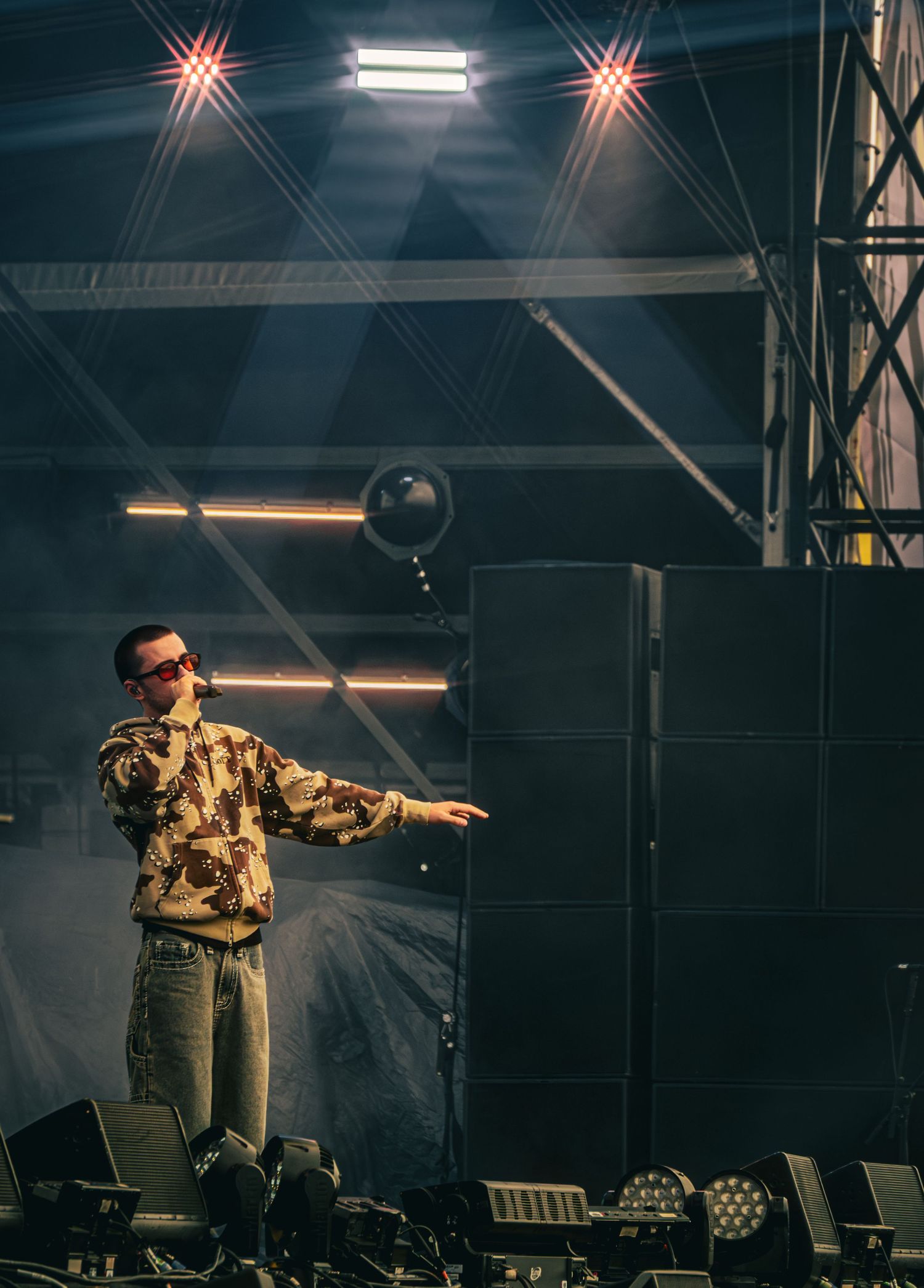 Performer in camo jacket and sunglasses on a lit stage with speaker stacks and a balloon floating overhead.