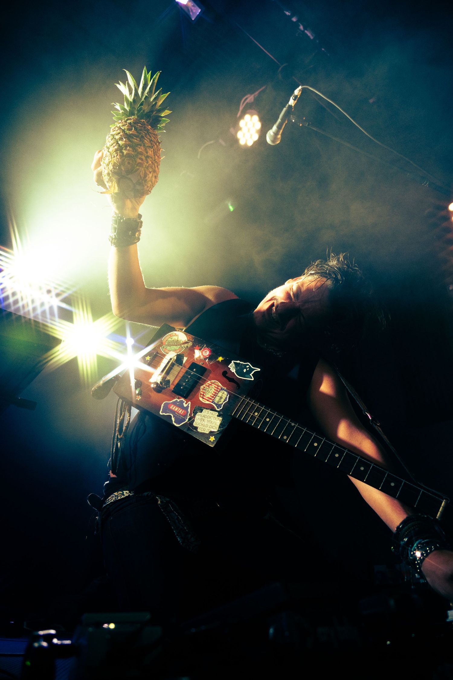 A performer on stage holds up a pineapple while playing guitar under dramatic stage lighting and smoke effects.