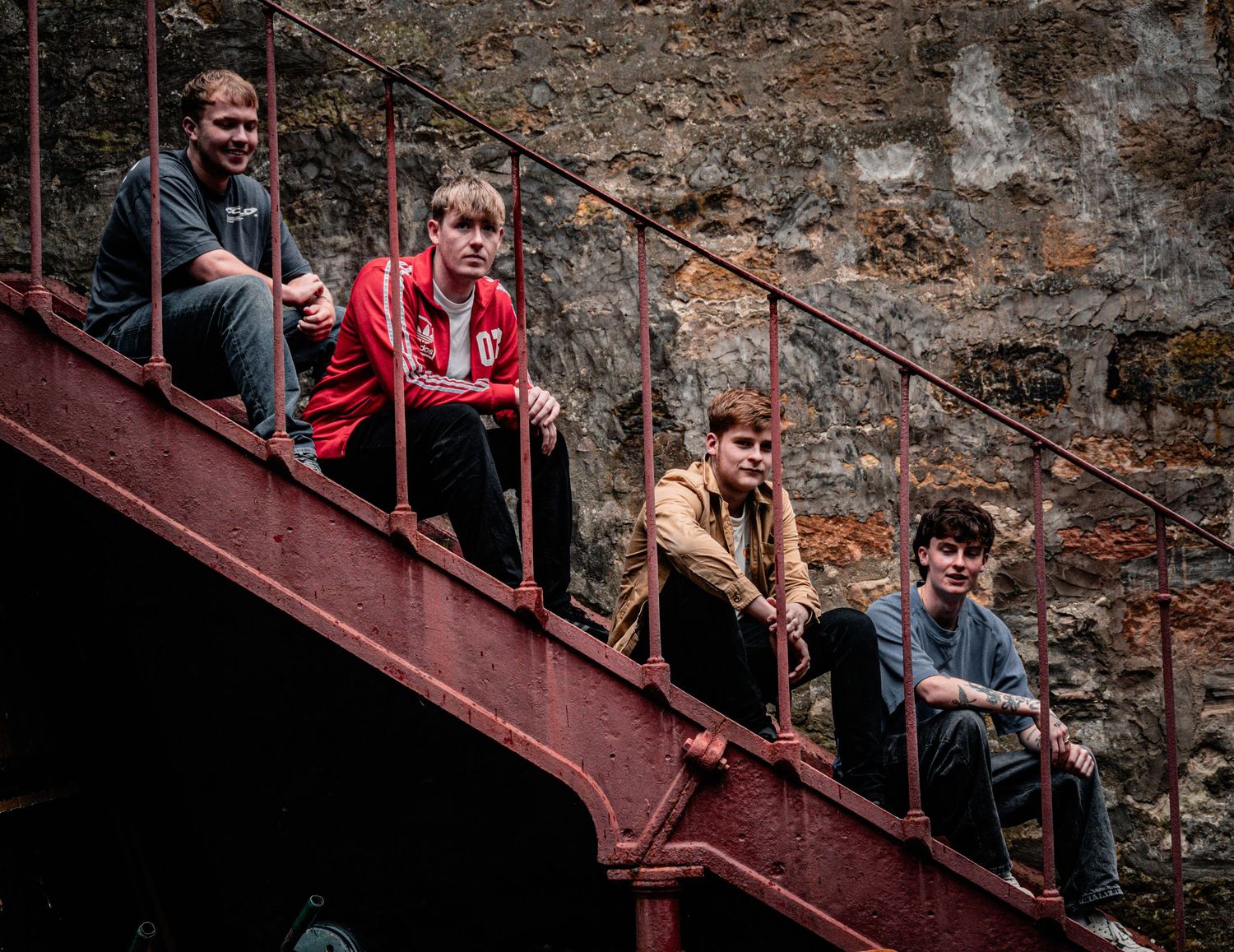 Four young men sitting on red metal stairs against a stone wall backdrop.