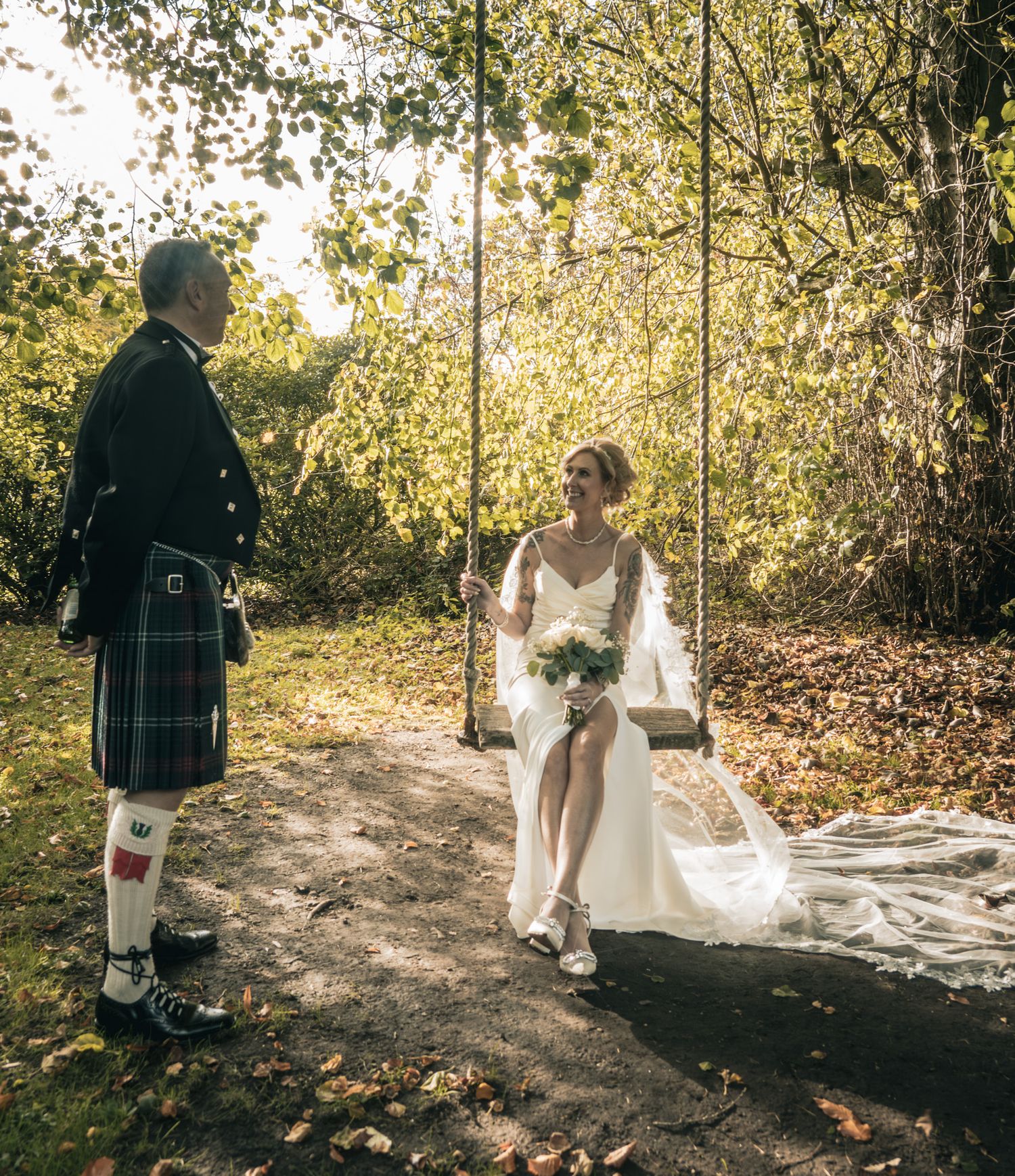 Bride in white gown sitting on a garden swing holding bouquet, groom in traditional Scottish kilt standing nearby.