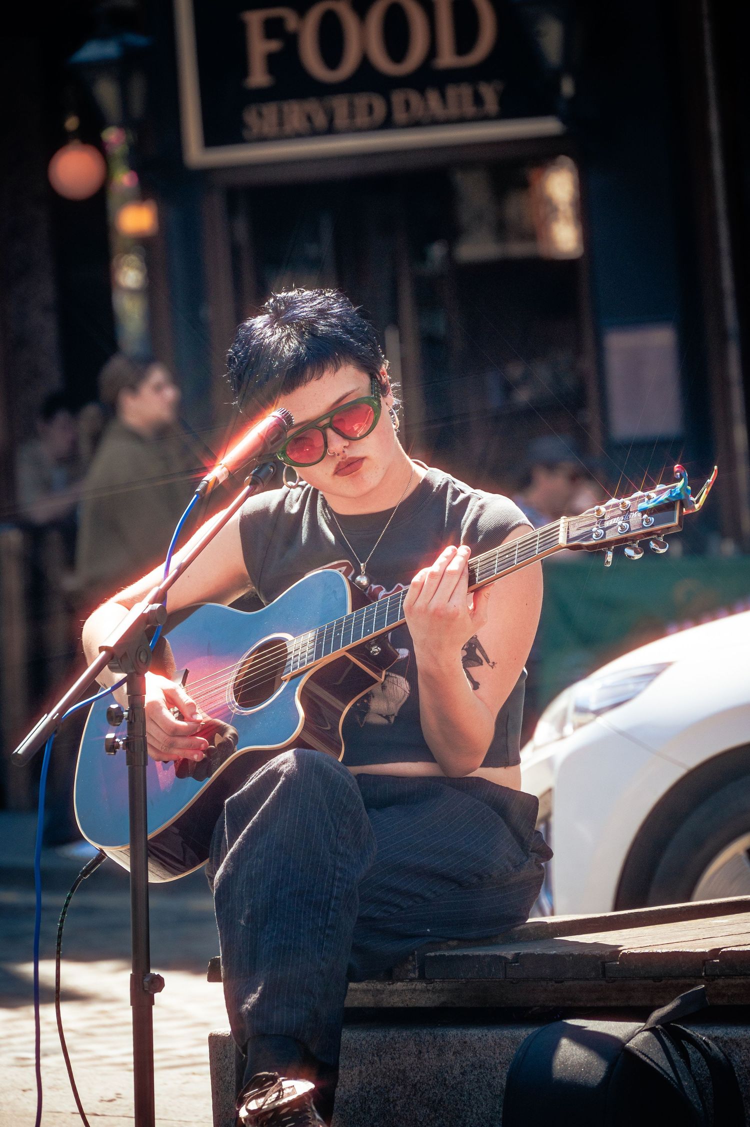 A young musician with short dark hair and red sunglasses plays acoustic guitar at an outdoor street performance near a food sign.