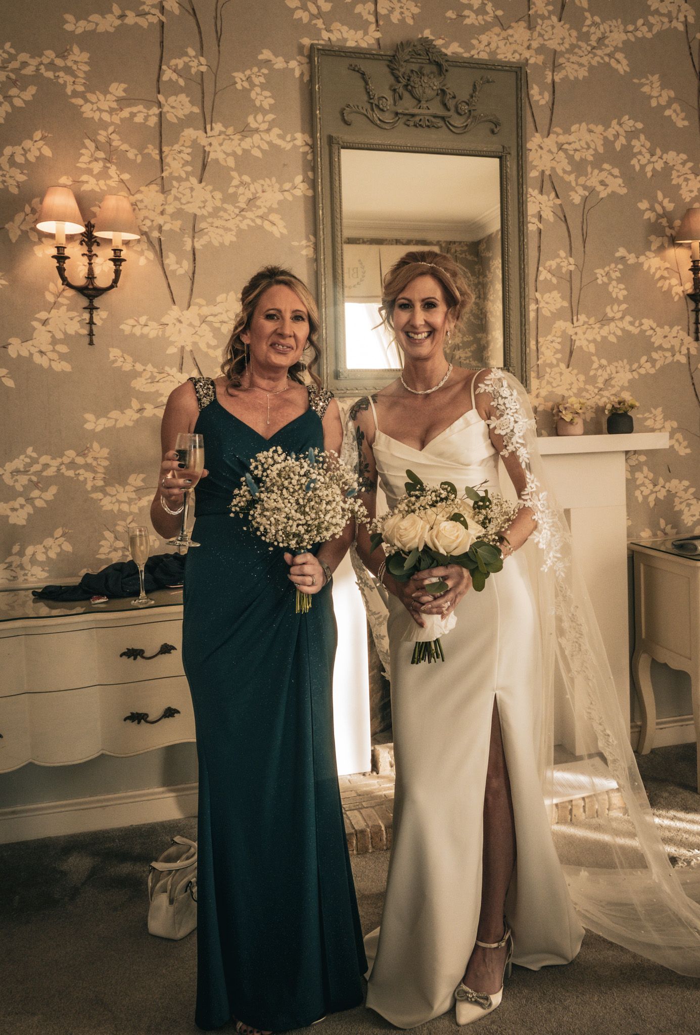 Bride in white lace gown holding roses bouquet poses with bridesmaid in teal dress holding champagne in elegant room.