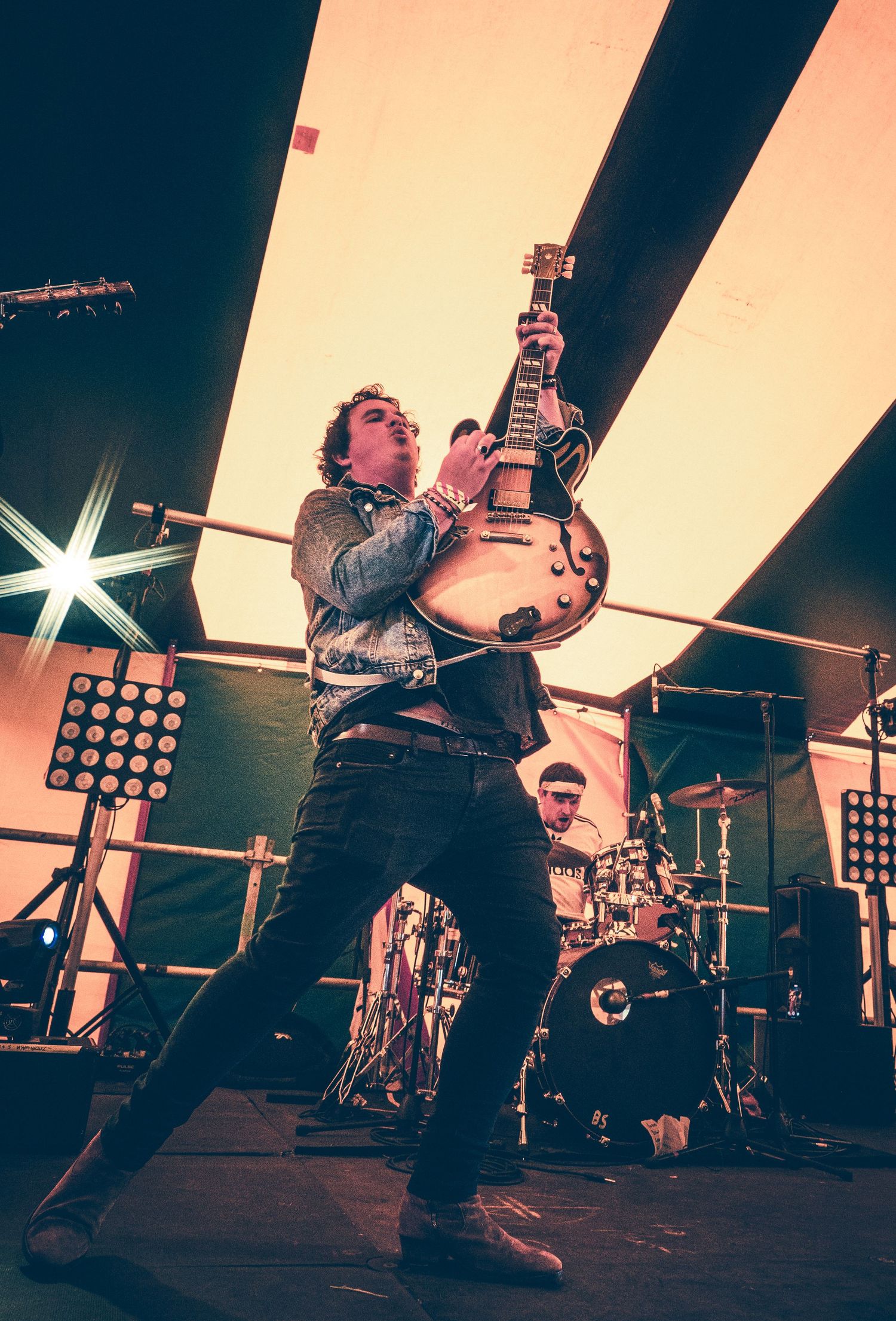 Rock guitarist playing electric guitar on stage with dramatic lighting and drummer in background.