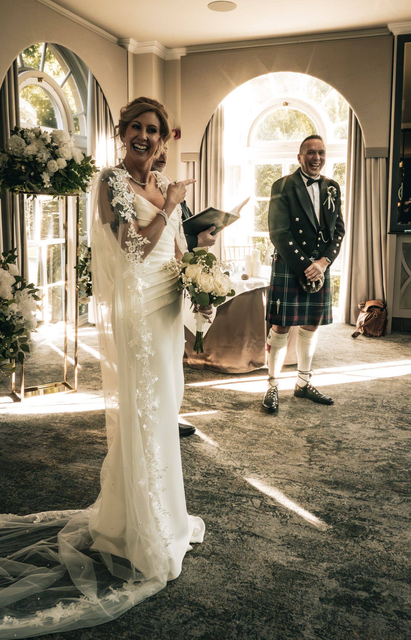 Smiling bride in lace wedding gown reads vows as groom in Scottish tartan kilt stands in doorway.