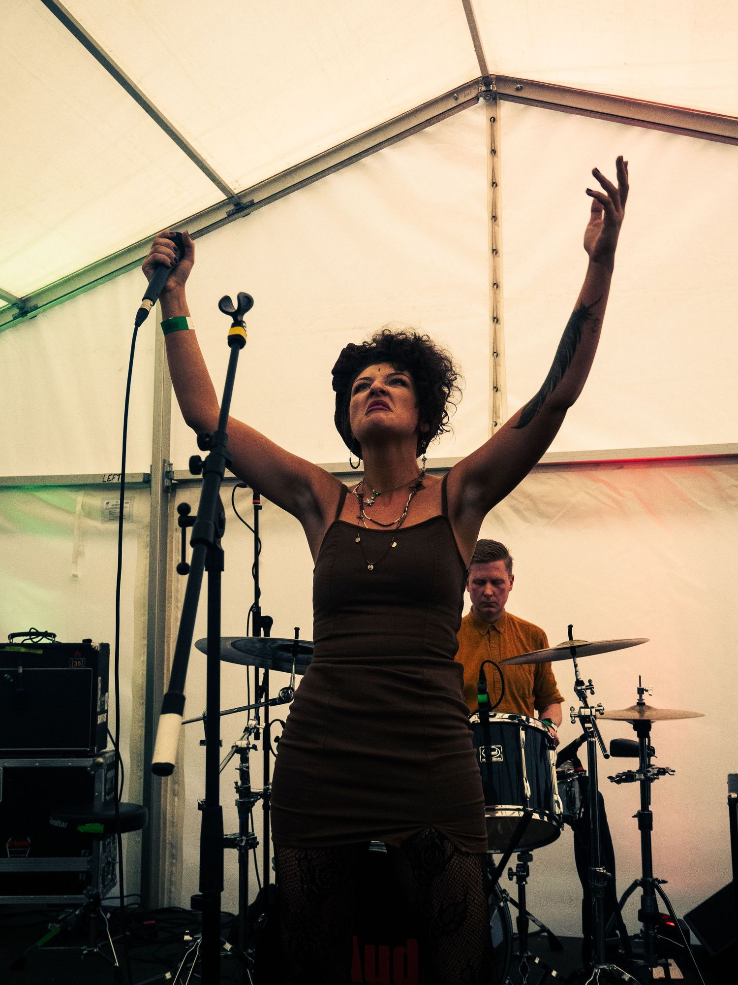 Singer performing with raised arms against tent backdrop with drummer visible in background.