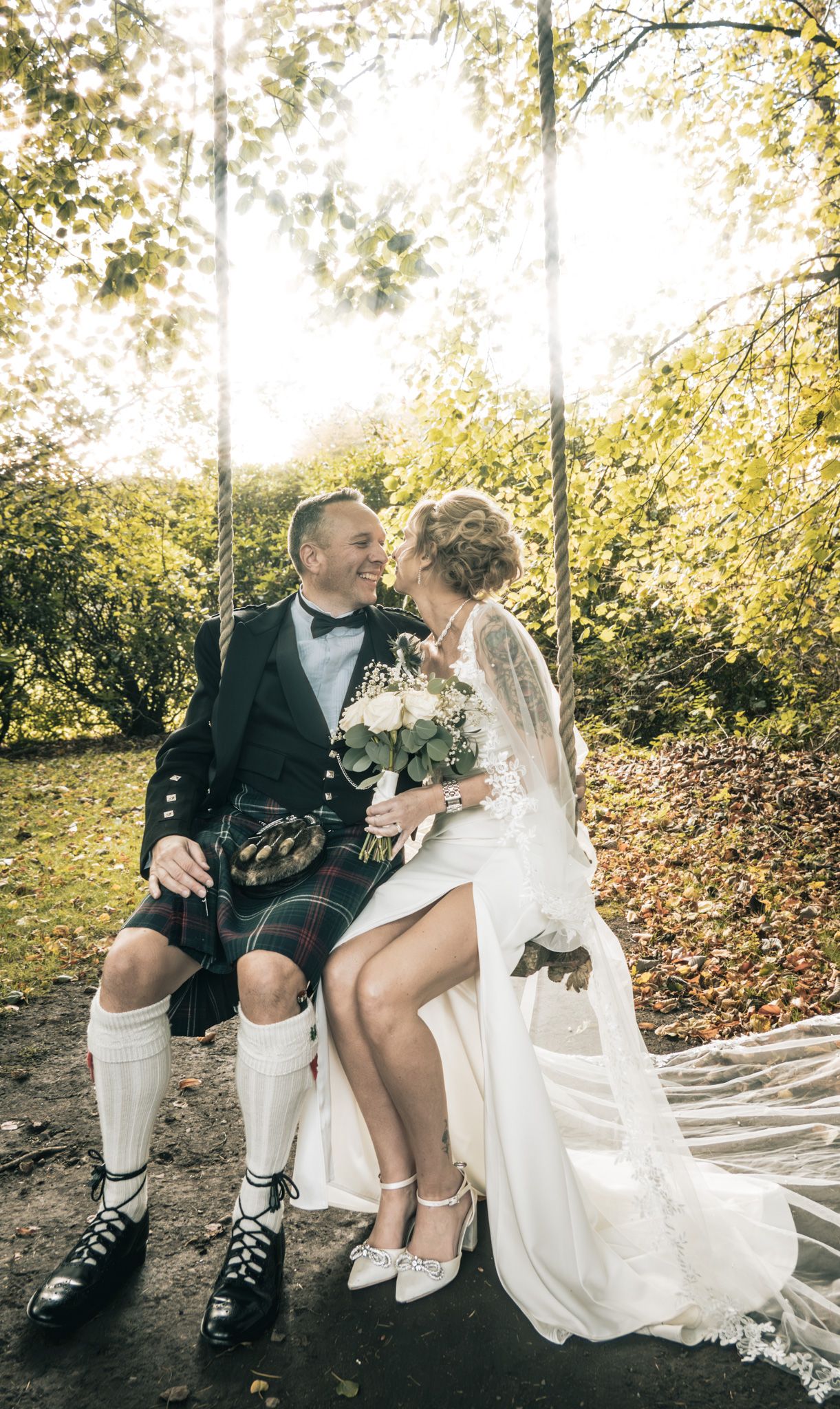 Bride and groom in Scottish kilt sit on swing in sunlit autumn garden, sharing a romantic moment.