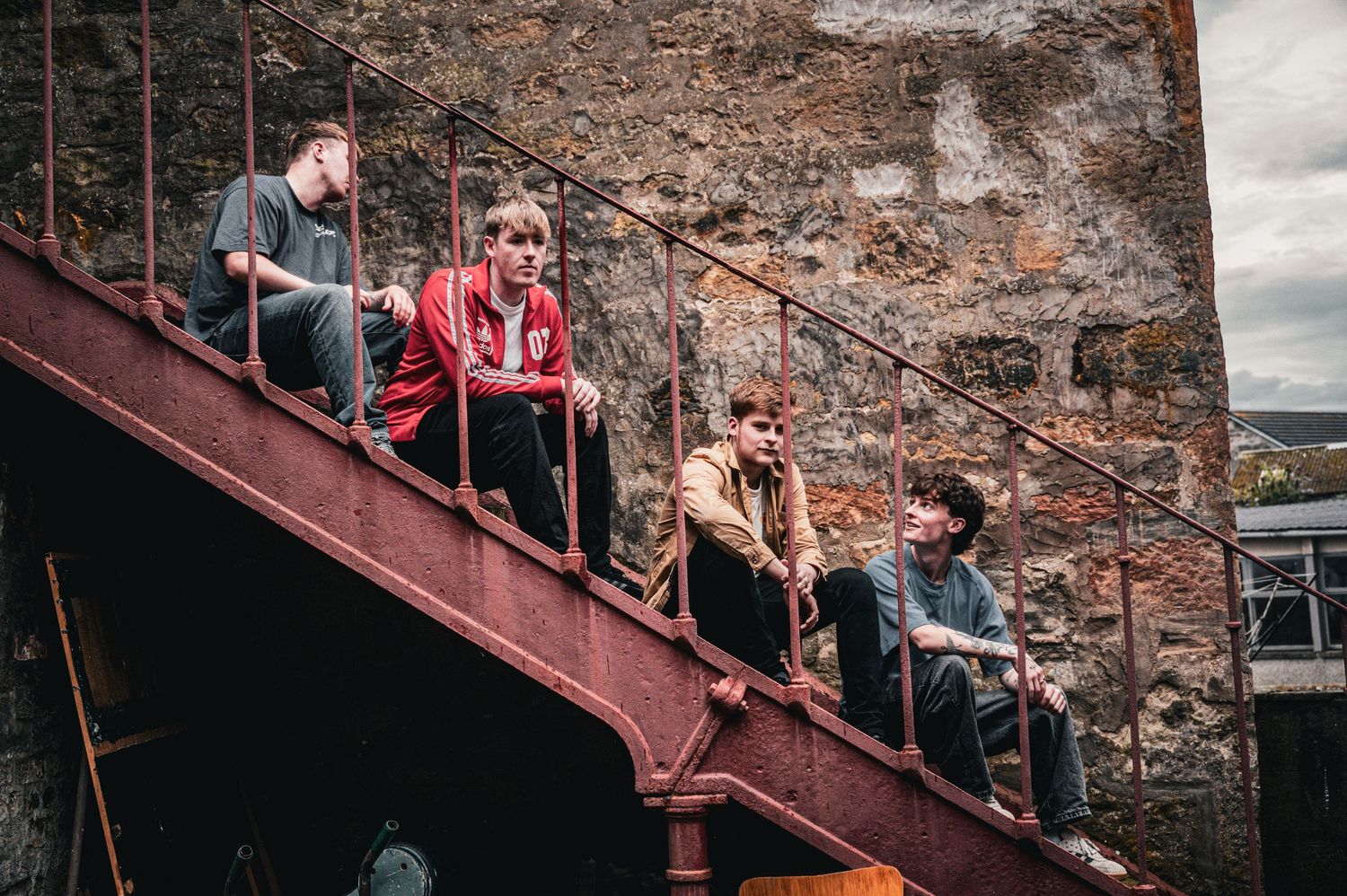 Four young men sitting on a rusty red metal fire escape staircase against a stone wall outdoors.