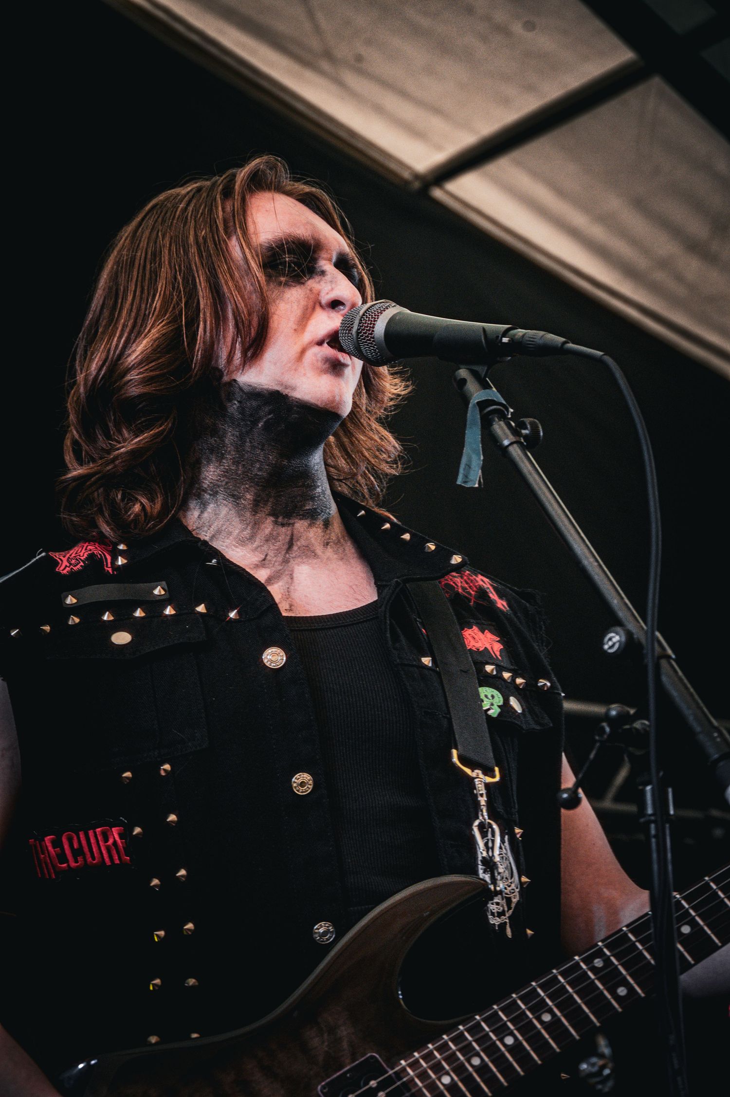Long-haired rock guitarist singing at outdoor stage wearing studded black sleeveless jacket.