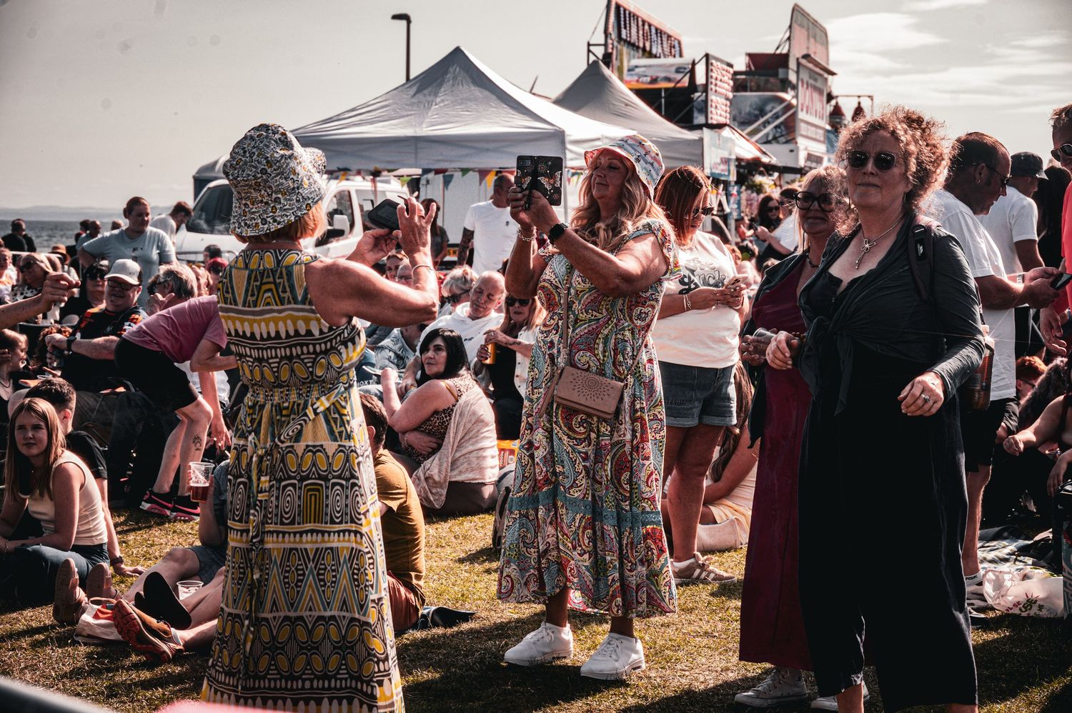 Crowd enjoying an outdoor music festival on a sunny day, with women dancing in colorful dresses on green grass.