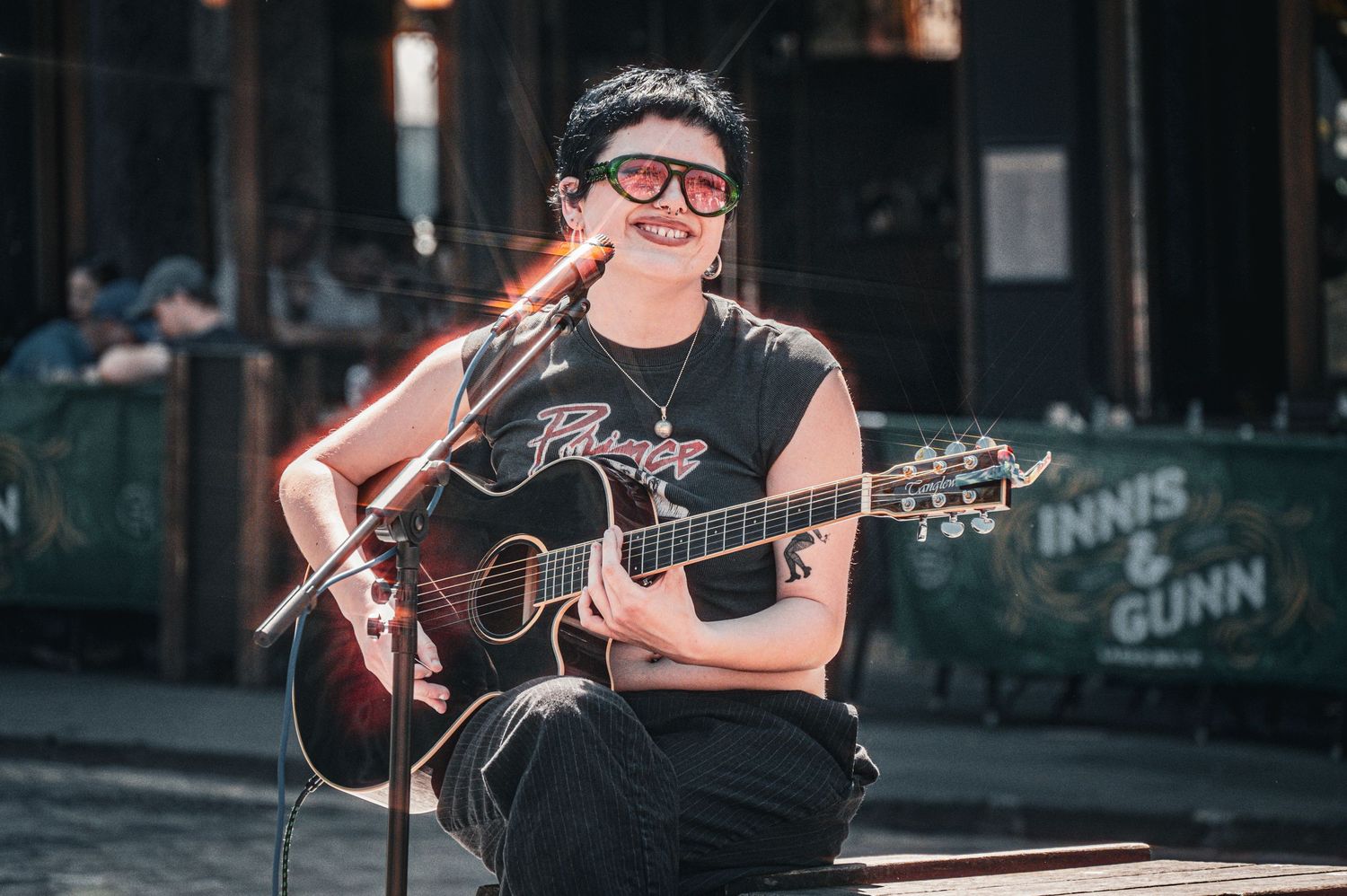 Smiling musician playing acoustic guitar outdoors, wearing black t-shirt and red glasses, performing near Innis & Gunn sign.