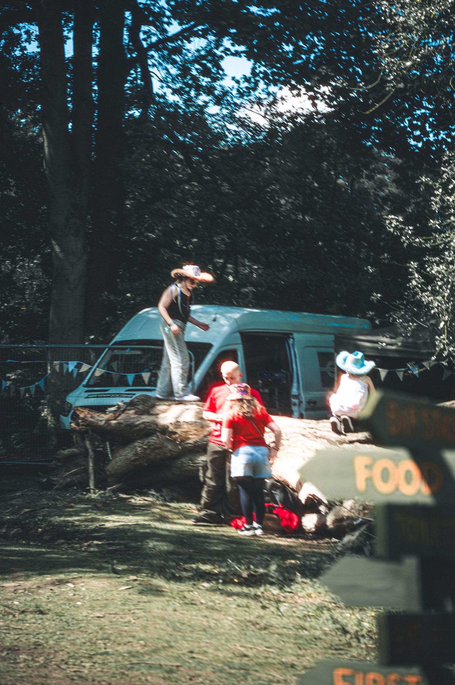 Children and adults gathered around fallen tree near vintage van at outdoor woodland festival.