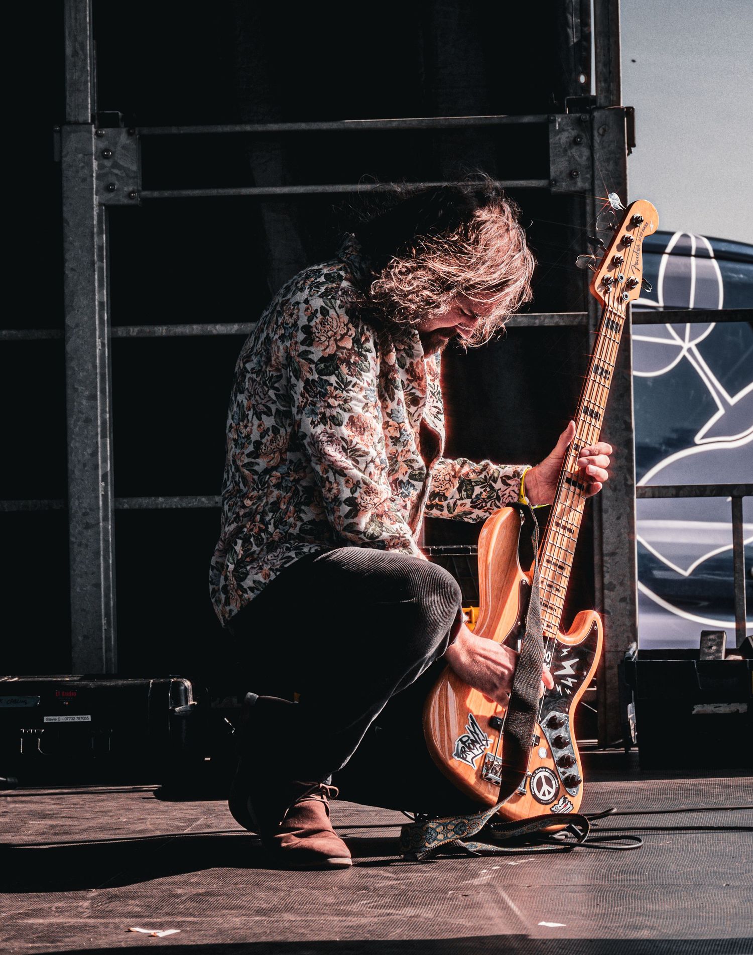Guitarist kneeling on stage, playing an orange electric guitar with stickers, wearing a floral shirt and jeans.