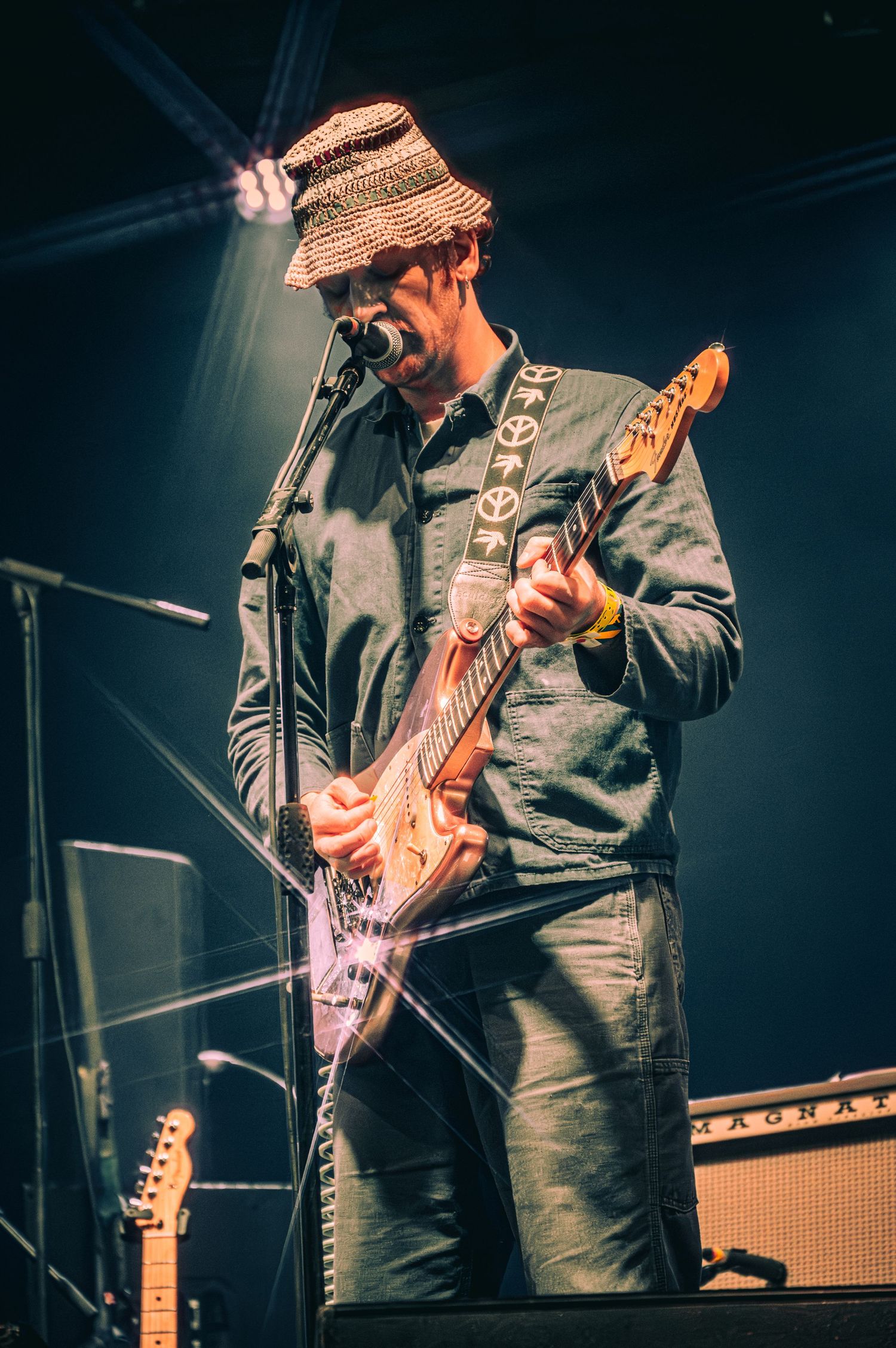 Musician playing electric guitar on stage wearing a straw hat and denim shirt under dramatic blue stage lighting.