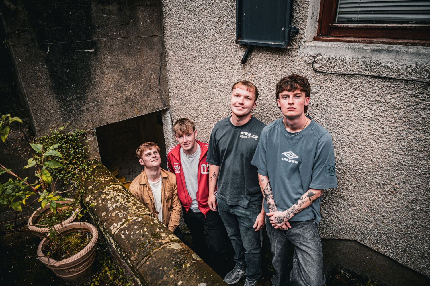 Four young men pose in a narrow alleyway between stone buildings, with plants and graffiti visible.