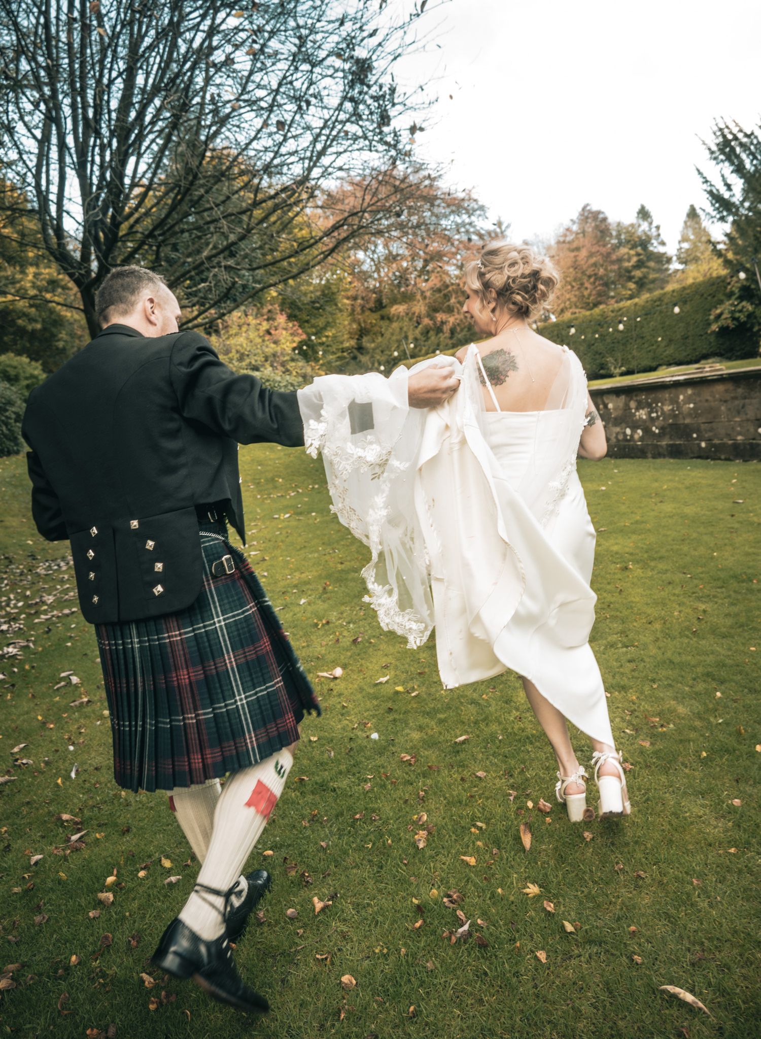 A groom in a traditional Scottish kilt holds his bride's wedding dress train as they walk across a lush green lawn.