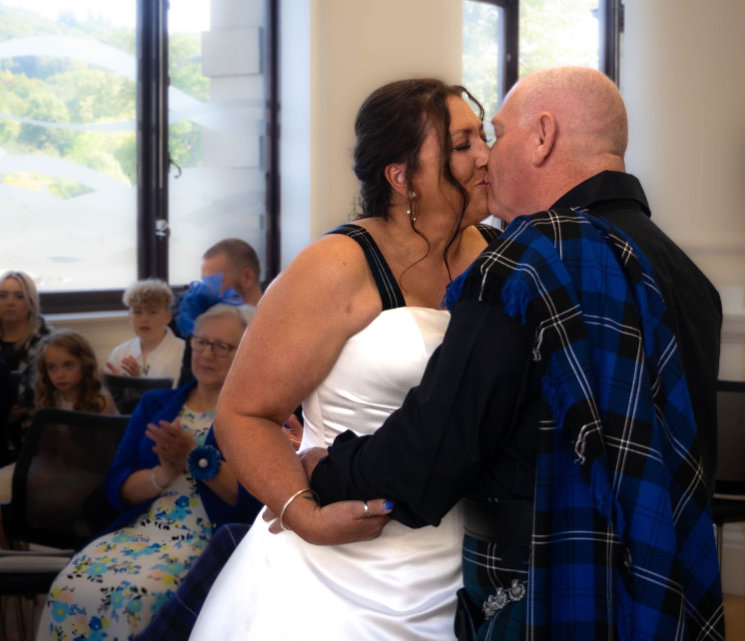 Bride and groom share a kiss at their wedding ceremony, with guests seated in the background.