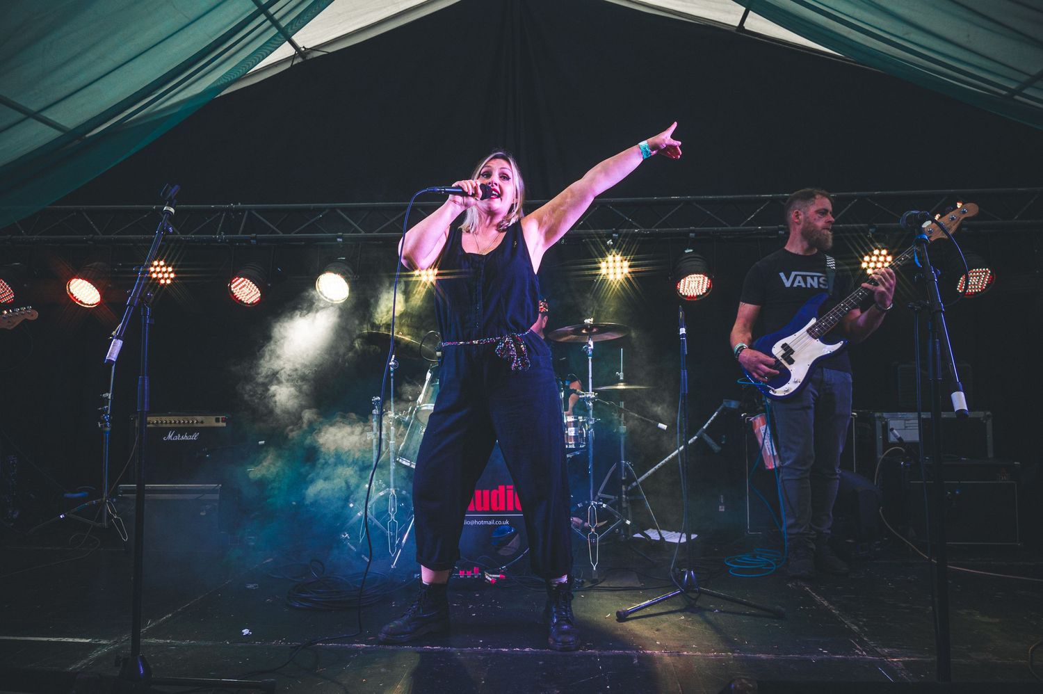 Rock band performing on stage with female vocalist pointing outward, guitarist, and dramatic stage lighting.