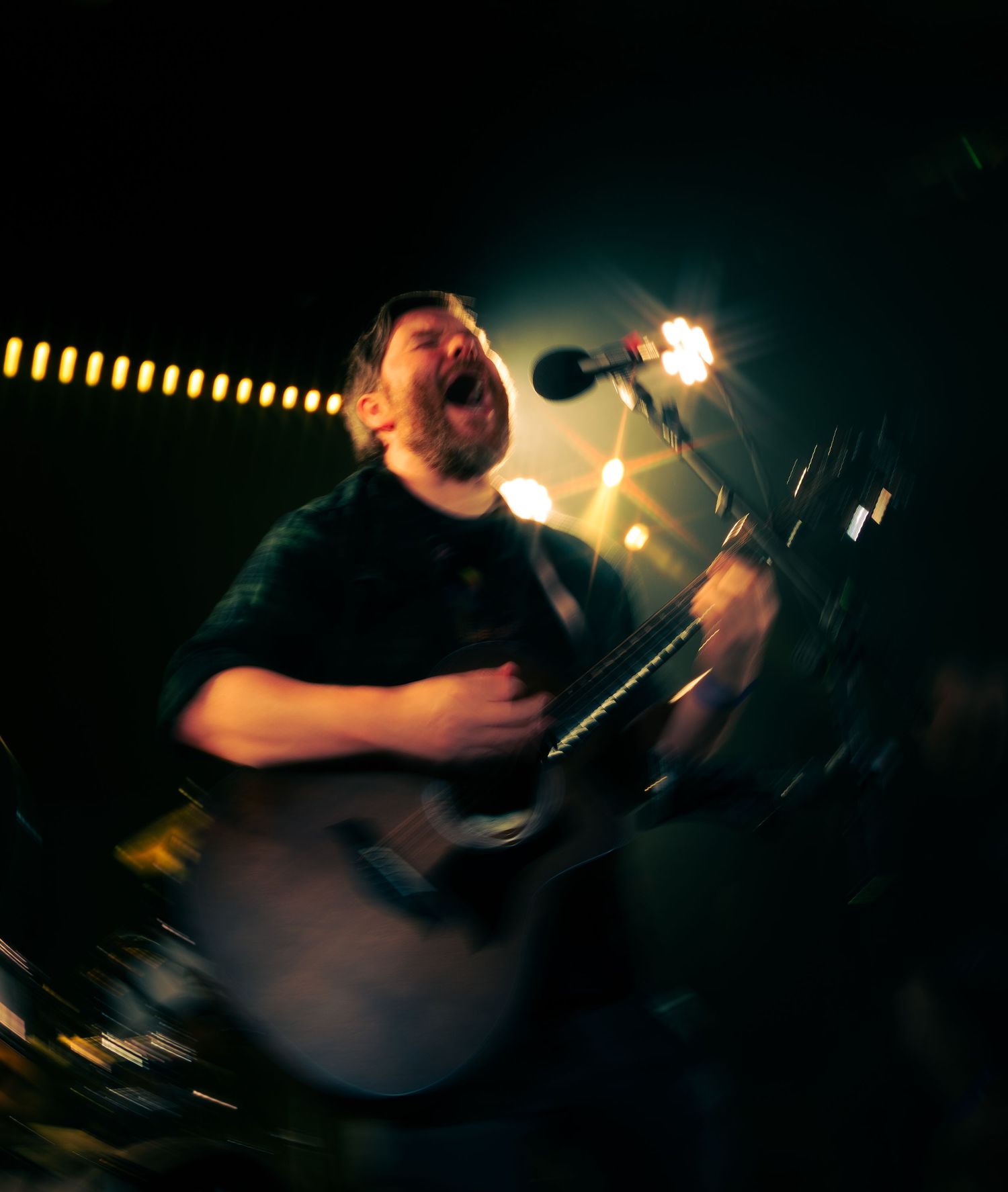 Musician performs passionately on acoustic guitar on dimly lit stage with string lights in background.