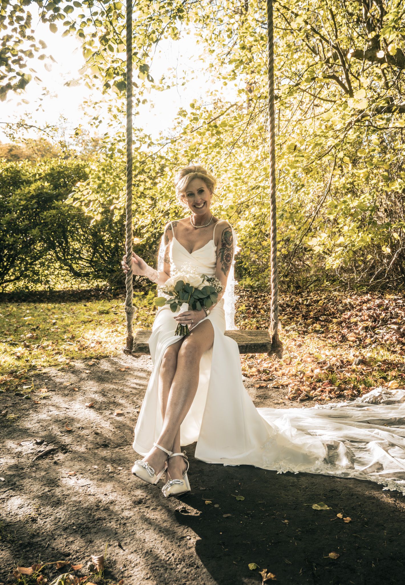Tattooed bride in white gown sits on garden swing holding bouquet in sunny outdoor wedding setting.