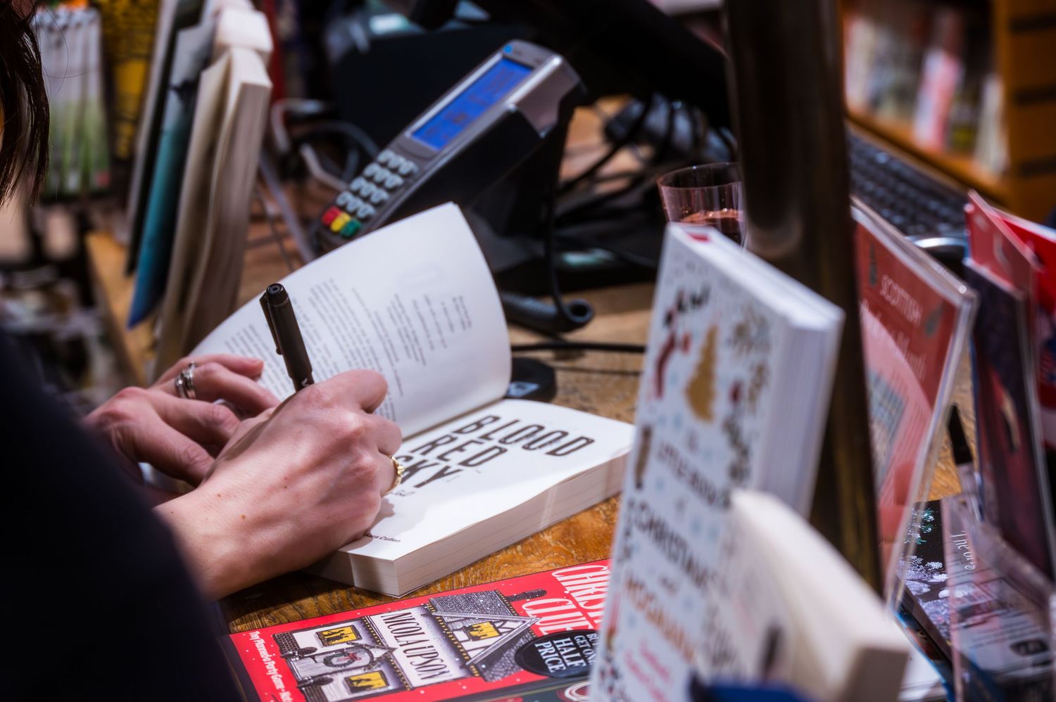 Author signing a book at a bookstore signing event surrounded by displayed books and a payment terminal.