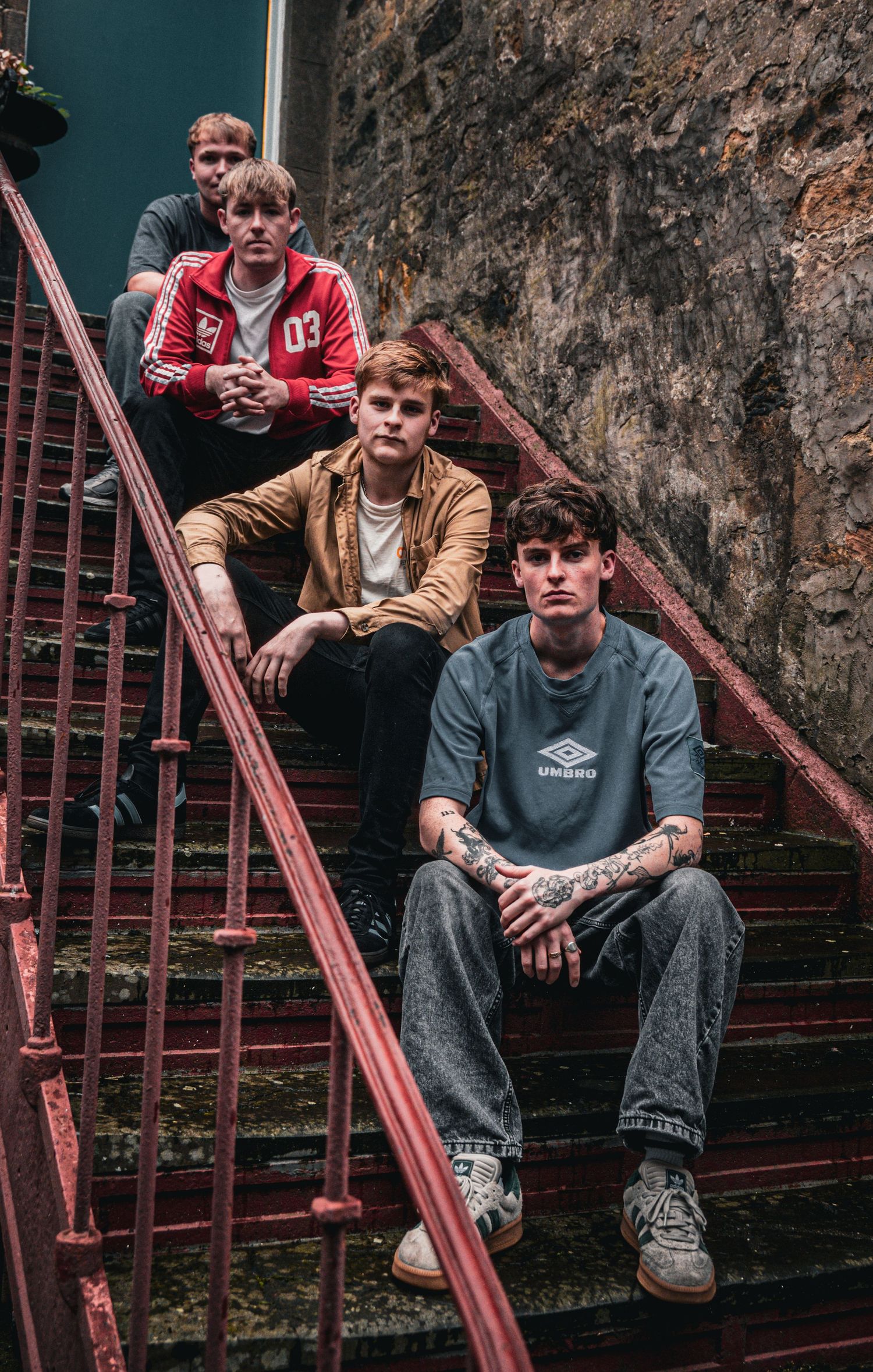 Four young men pose on red metal stairs against a stone wall, one with tattoos wearing an Umbro shirt.