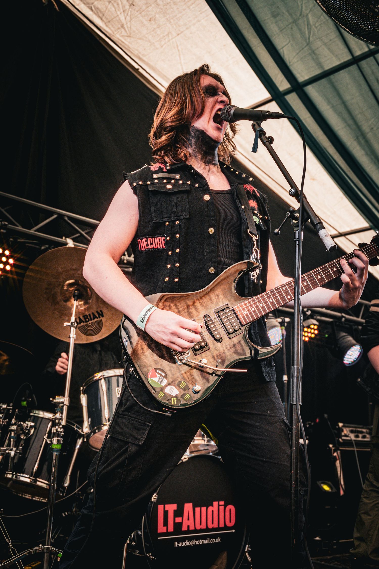 Rock guitarist performs on stage with electric guitar and microphone, wearing black vest and jeans at LT-Audio event.