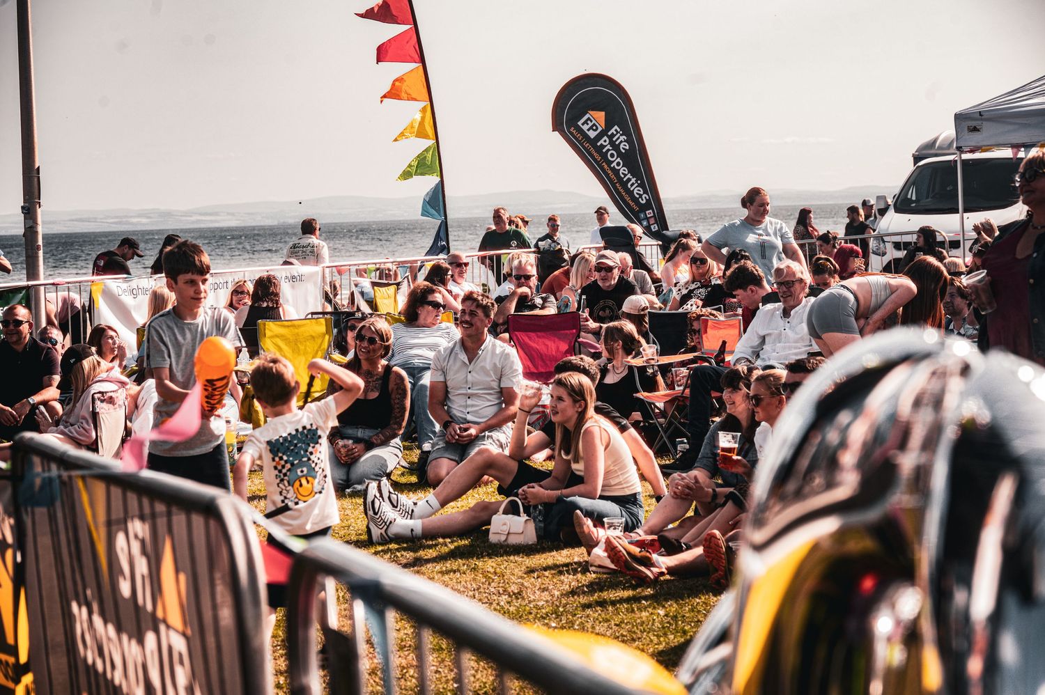 Crowd of people enjoying an outdoor beach festival with colorful flags and a sea view on a sunny day.