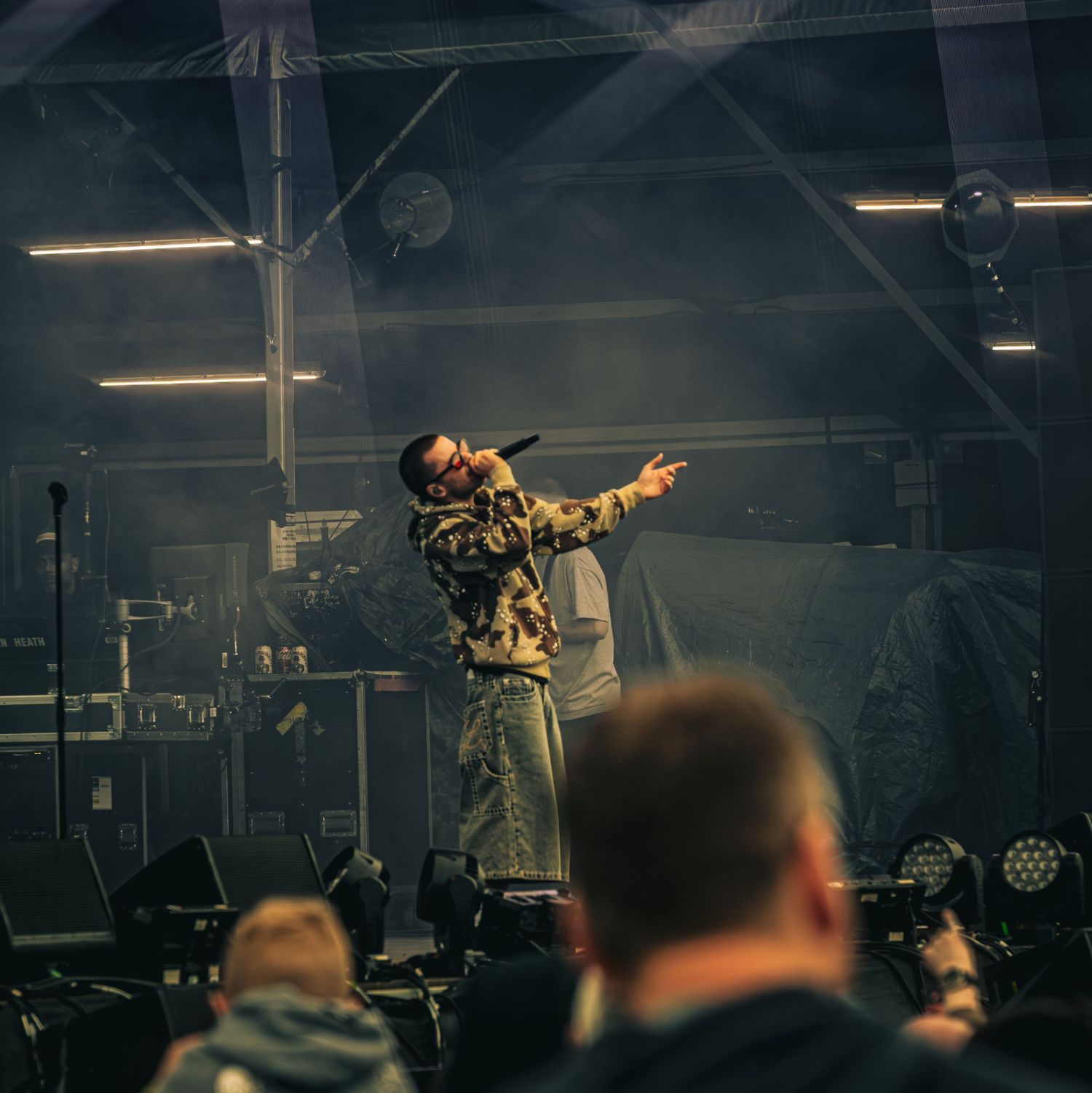 A rapper performing on stage in camouflage outfit, pointing at crowd in a dimly lit indoor venue.
