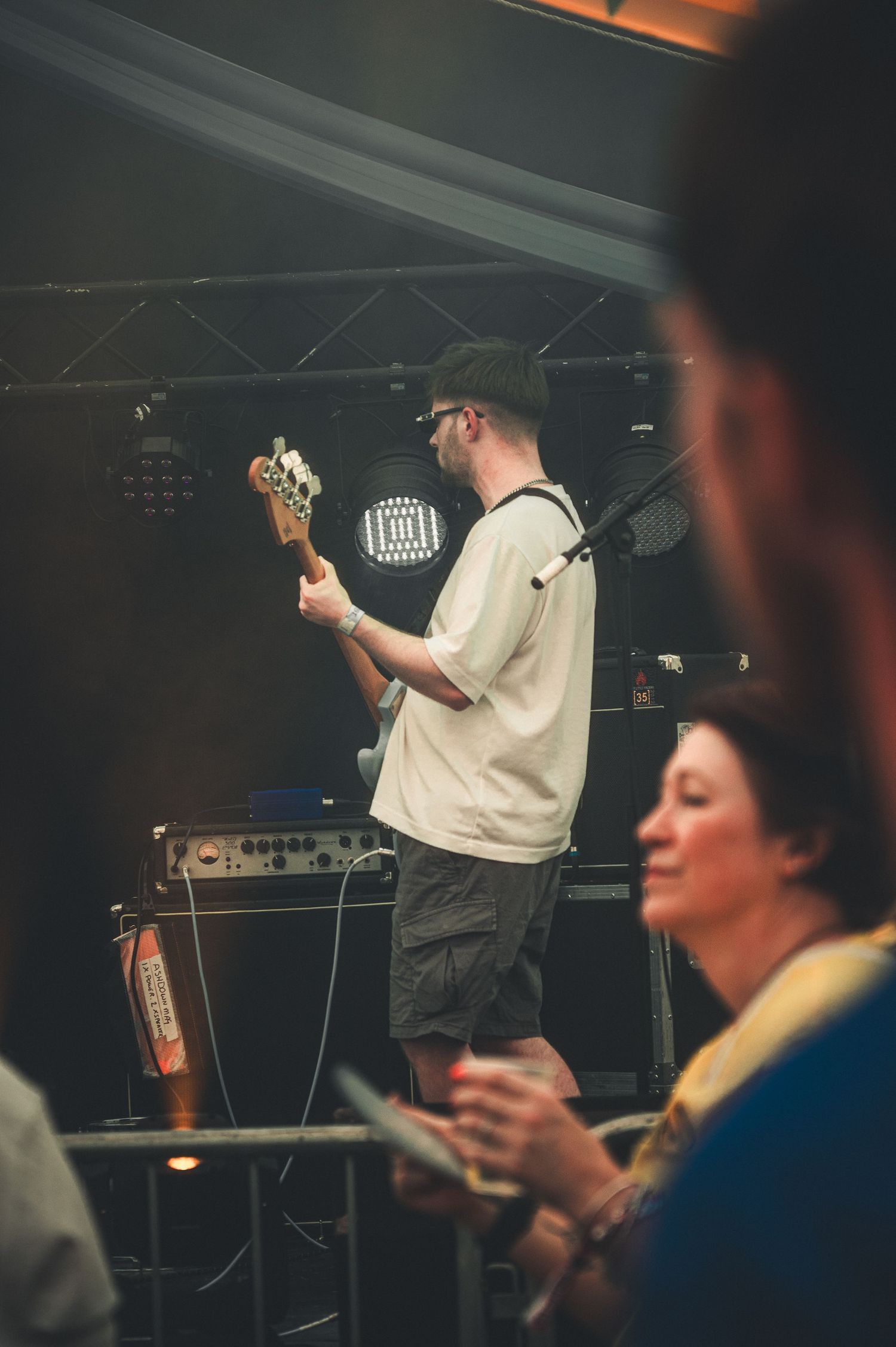 A guitarist performing on stage in a white t-shirt and cap, with audience members in the foreground at a live music event.