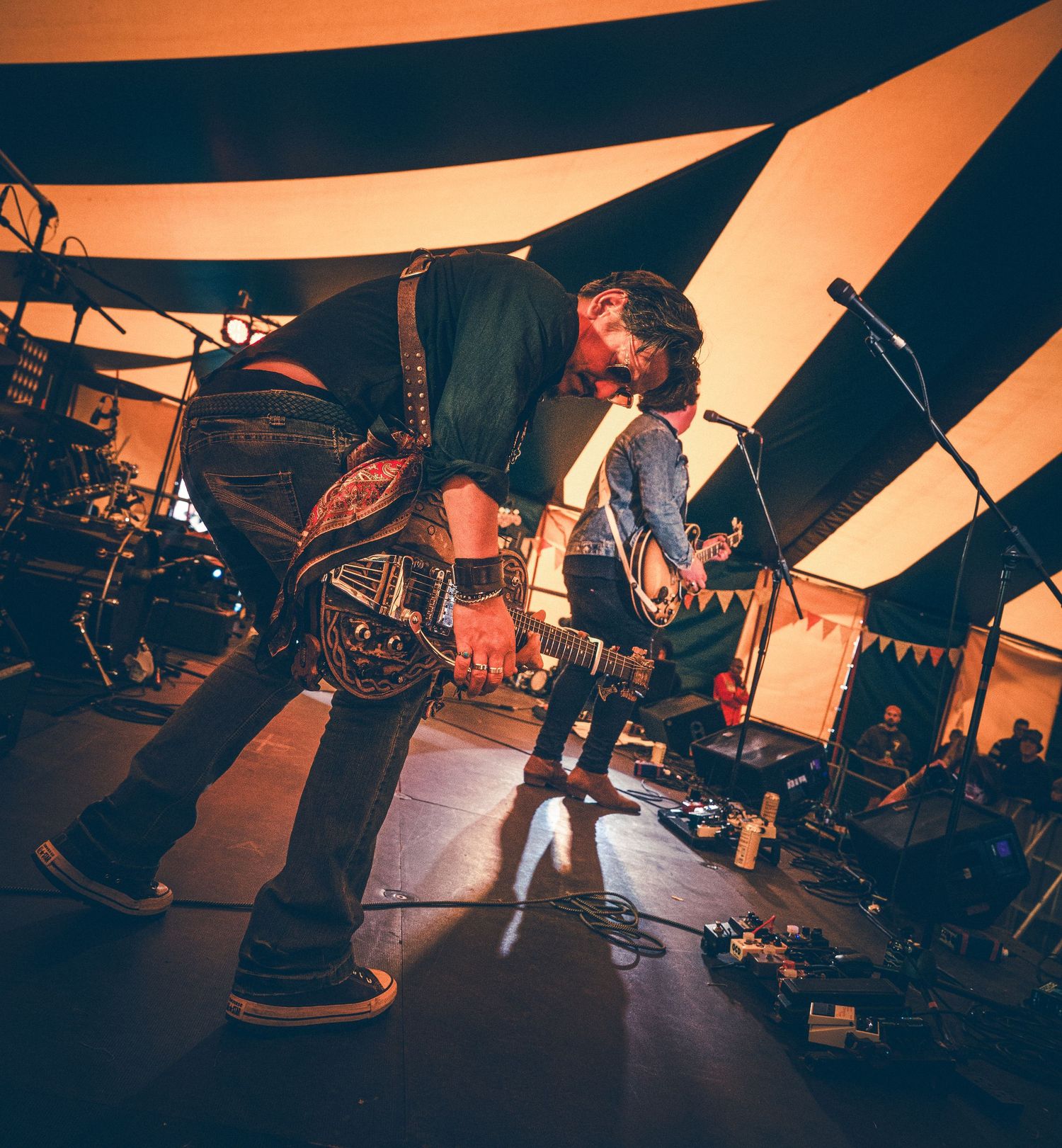 Two guitarists perform energetically on stage under a striped tent at an outdoor music festival.