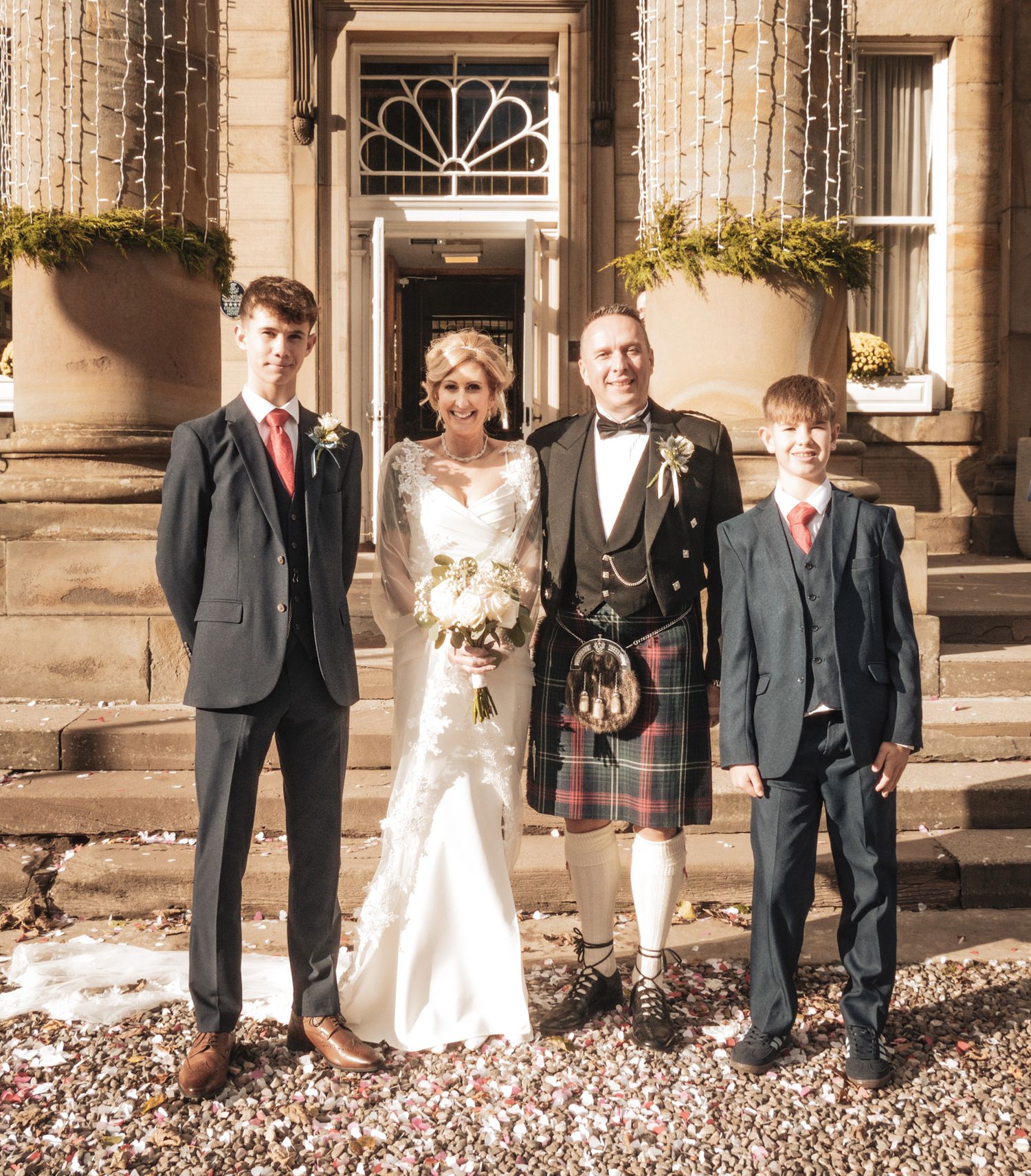 Wedding party posing outside a Scottish manor, groom in tartan kilt with bride in white gown flanked by two young men in suits.