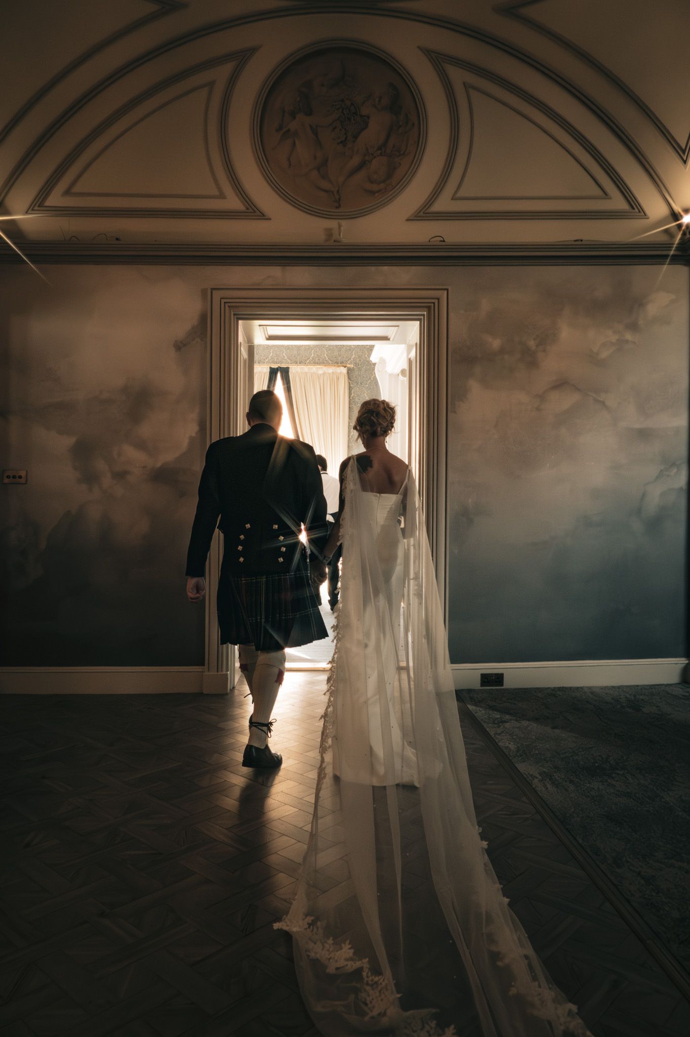 Bride in white gown and groom in Scottish kilt walk through ornate doorway in dimly lit wedding venue.